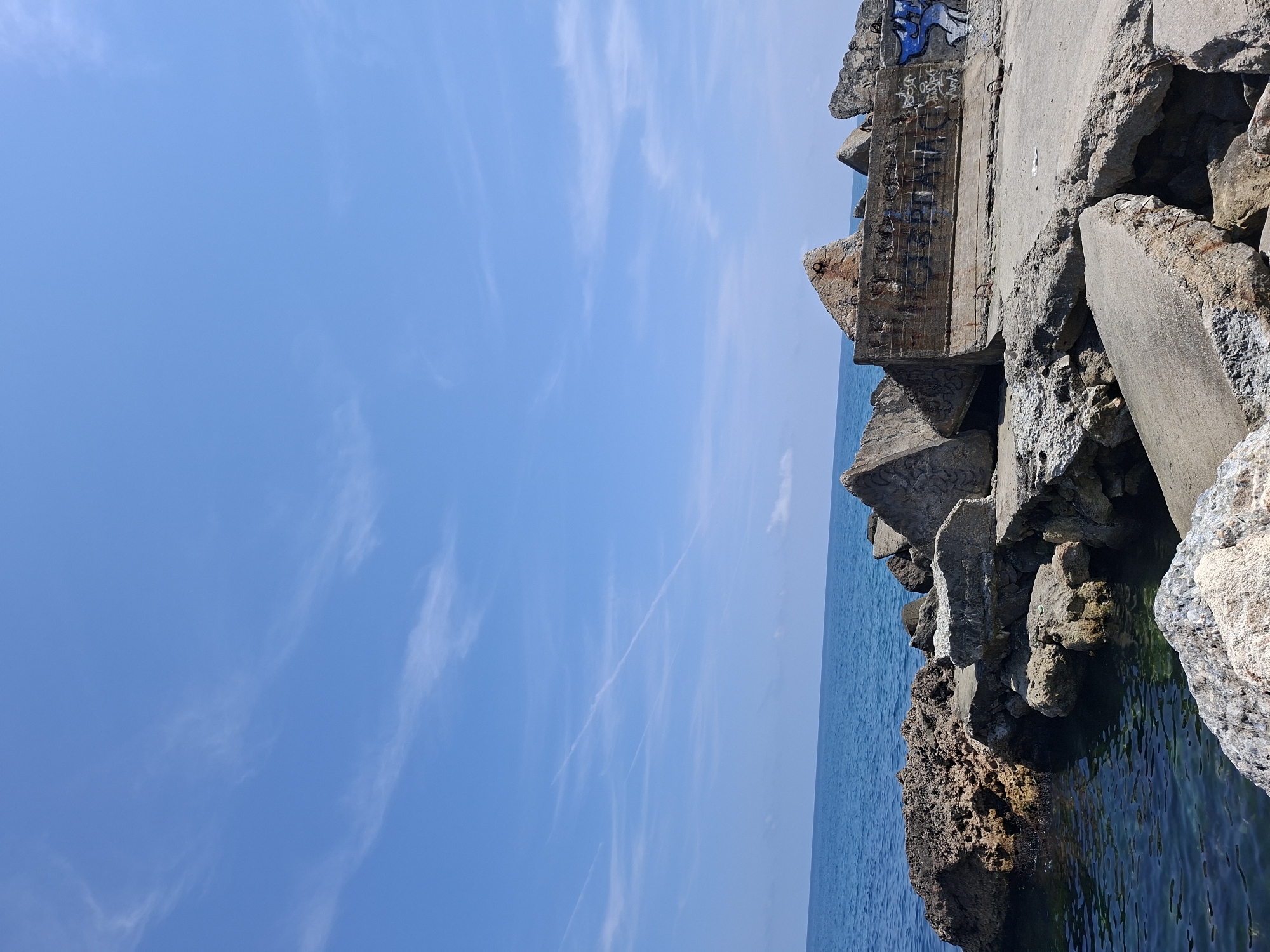 The image shows a seascape with a breakwater constructed of concrete and rocks. The breakwater is situated in Balgarevo, Bulgaria. The sea is calm and a clear blue, with a horizon visible in the distance. The sky is also blue with thin, wispy clouds and contrails from an aircraft. The concrete structures are weathered and show signs of graffiti. The rocks are of various sizes and shapes, some submerged in the water, others forming part of the breakwater. There are no people or animals visible in the scene. The overall impression is one of a coastal defense structure in a natural environment.