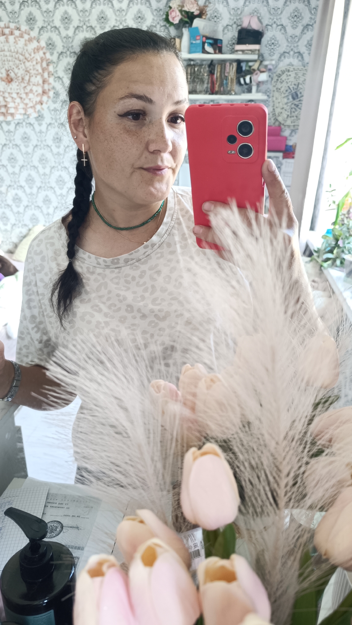 A medium shot captures a woman taking a selfie in a room, likely her home, in Chaikovsky, Russia. She is positioned in the center, holding a red smartphone up to take the photo, with her face partially obscured by her hand and the phone. She has dark hair pulled back into a braid and is wearing a light-colored shirt with a leopard-print pattern. She has freckles across her nose and cheeks and is wearing a green beaded necklace. In the foreground, a bouquet of light pink tulips and fluffy white plumes obscures some of the lower portion of the image. Behind her, shelves filled with various items, including what appear to be decorative objects and personal belongings, are visible against a wallpapered wall. A window is present to the right, letting in natural light. The overall impression is of a casual, personal moment captured in an indoor domestic setting.