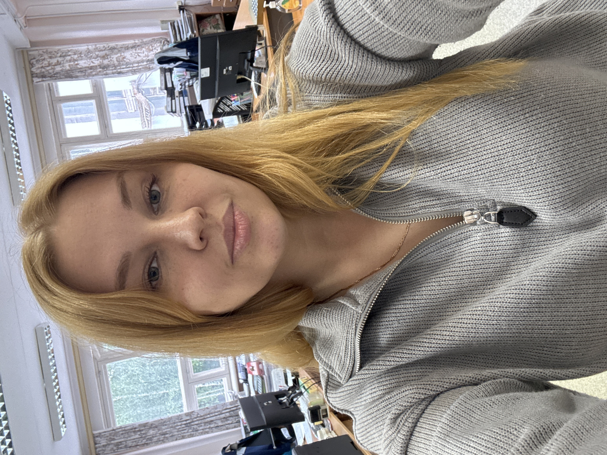 A selfie of a fair-skinned person with long, light brown hair is presented. The person is wearing a gray, knitted, zip-up sweater. A thin gold necklace is visible around their neck. The background shows a window with a light-colored curtain. A desk with office equipment, including a monitor and files, is partially visible. The location is possibly an office setting within the city of Unknown, Unknown.