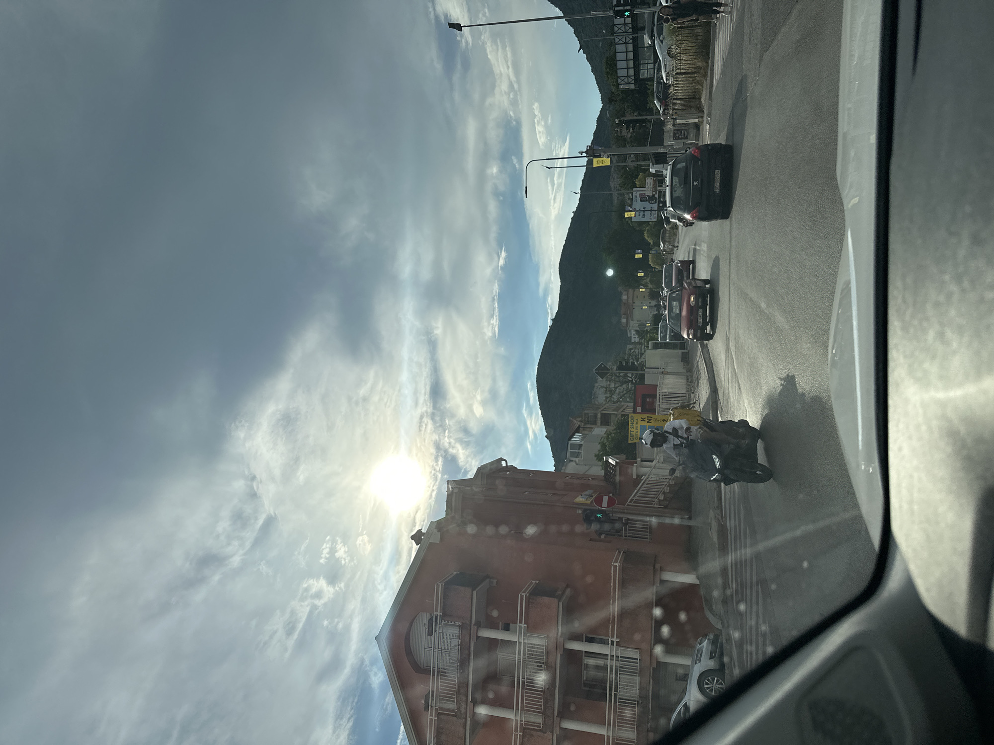 The image depicts a street scene in Budva, Montenegro, viewed from inside a vehicle. The sun is shining brightly through scattered clouds, casting a warm light on the surroundings. On the left side of the road, a four-story building with a reddish-orange facade features multiple balconies with white railings. A traffic light is visible, showing a green signal, next to a red and white circular sign. A person on a dark-colored scooter, wearing a white helmet and a white jacket with a yellow backpack, is riding on the road. Several cars are also on the street, including a red hatchback, a black Peugeot, and a silver sedan parked on the left. In the background, green hills rise, and streetlights are present along the road. The road surface is asphalt, and pedestrian crossing lines are visible. The overall environment suggests a sunny day in an urban area with mountainous terrain nearby.
