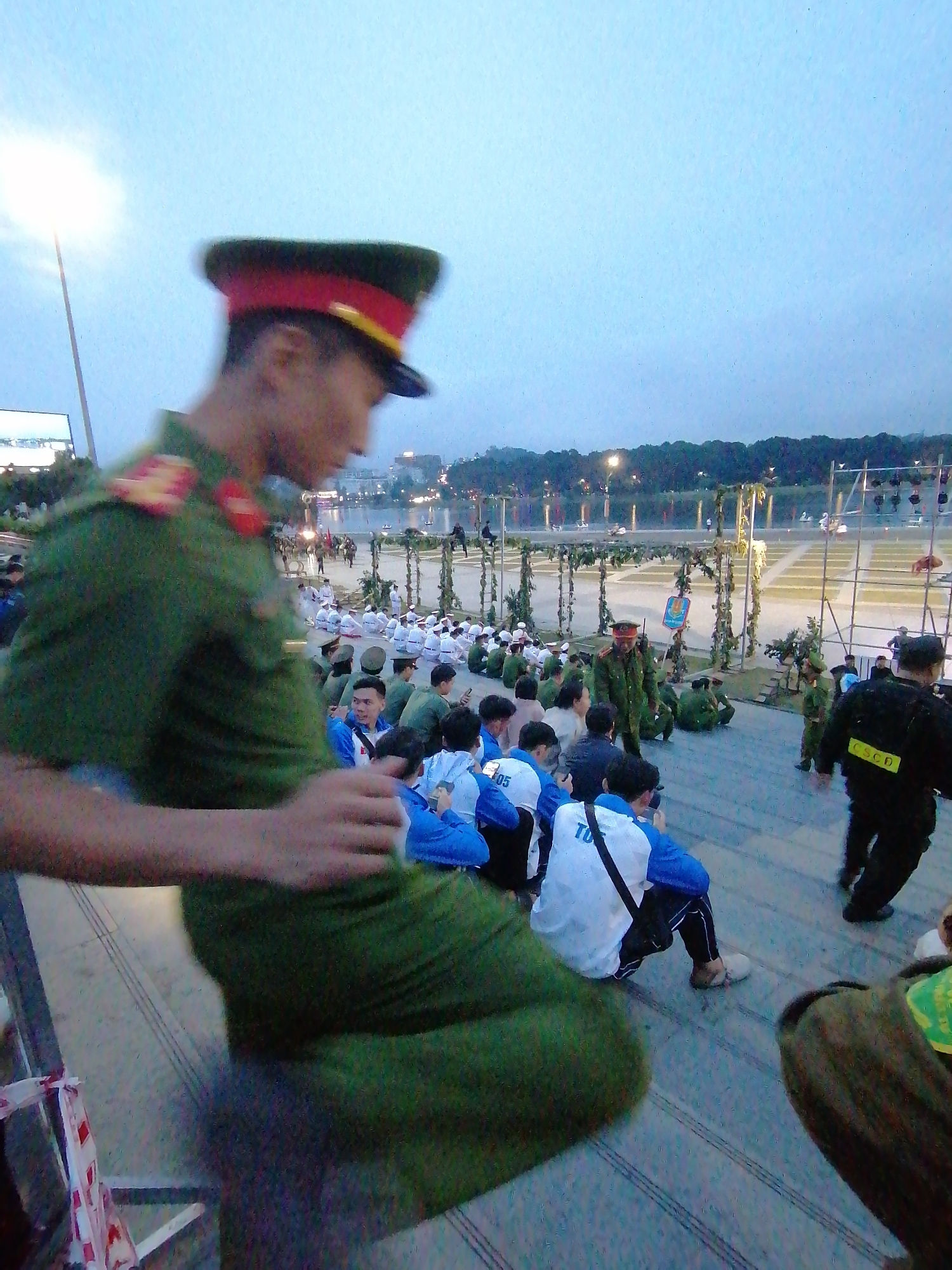 In Da Lat, Vietnam, a group of people are gathered outdoors, likely for an event. The scene appears to be set in the late afternoon or early evening, with soft, diffused light and a hazy sky suggesting either dusk or overcast conditions. Many individuals are seated on the ground, some in uniforms and others in casual attire.
In the foreground, a person in a green military-style uniform with a red and yellow cap is partially visible, facing away from the camera and seemingly interacting with someone or observing the scene. Their uniform is detailed with red insignia on the shoulders. Behind them, a crowd of people are seated in rows. Many of these individuals are wearing blue and white jackets, some with visible text on the back.
Further back, more individuals in green uniforms are present, along with a person in a black uniform with yellow lettering on the back that reads "CSCD". The background reveals a body of water, possibly a lake, lined with trees. Structures that appear to be stages or platforms are set up near the water, decorated with greenery, and illuminated by lights. The overall atmosphere seems orderly, possibly a public gathering, performance, or ceremony. The people's postures suggest they are attentive, with no strong indicators of distress or excitement.