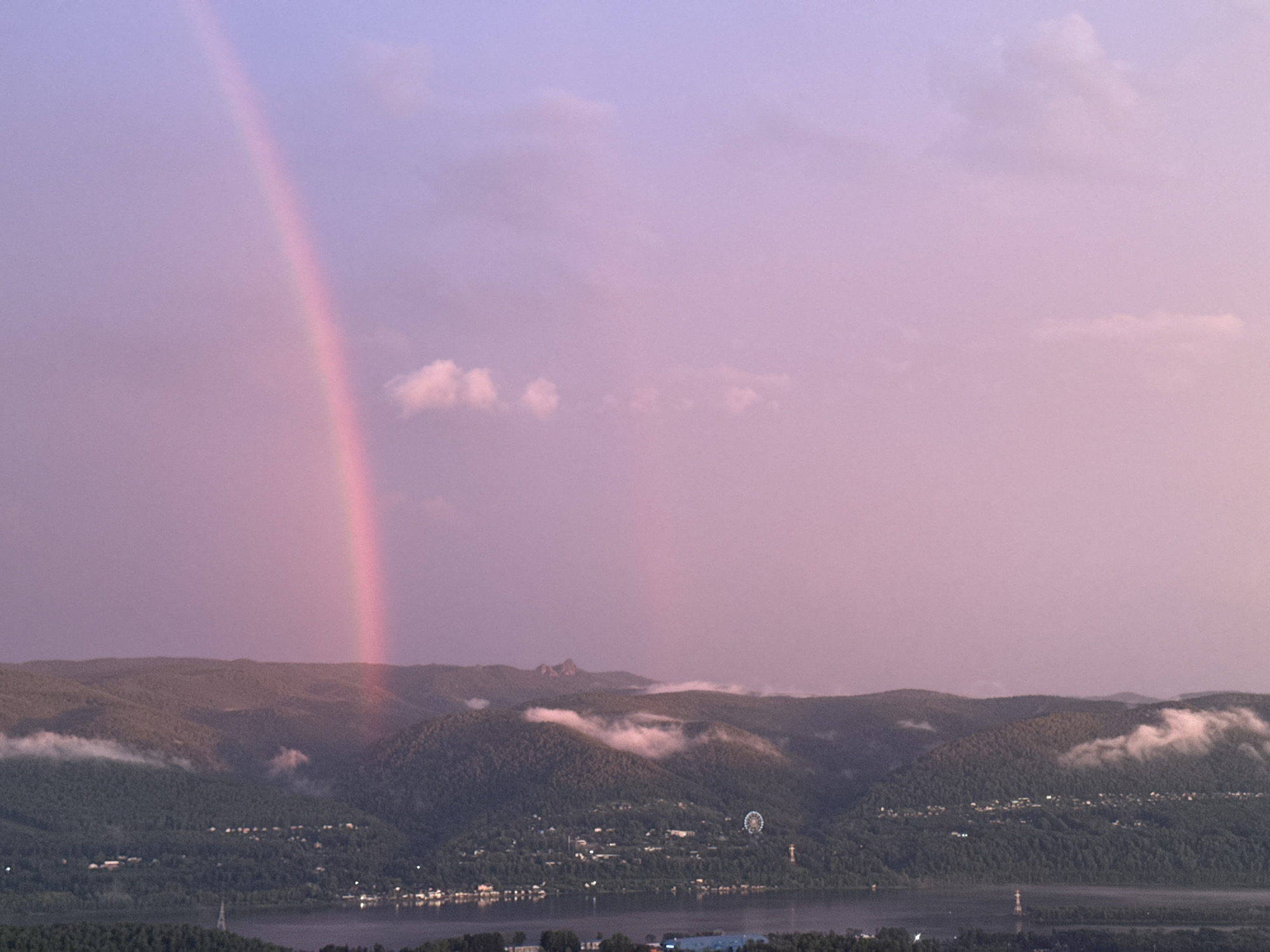 The image depicts a panoramic view of the city of Krasnoyarsk, Russia, during a sunset with a prominent rainbow. The foreground features a wide river, likely the Yenisei River, reflecting the soft, twilight sky. Beyond the river, rolling, tree-covered hills form the middle ground. Clustered settlements and scattered lights are visible on the slopes of these hills, indicating the urban presence within the natural landscape. A Ferris wheel is discernible on one of the hills, a recognizable landmark in the city. Wisps of clouds hover around the peaks of the hills, adding a dramatic effect. In the upper portion of the image, the sky transitions from a soft pink and lavender hue near the horizon to a lighter purple and pale blue higher up. A vibrant pink rainbow arcs across the sky on the left side of the frame, with a fainter secondary rainbow visible to its right. The overall scene is serene and picturesque, capturing the natural beauty of Krasnoyarsk bathed in the atmospheric light of sunset.