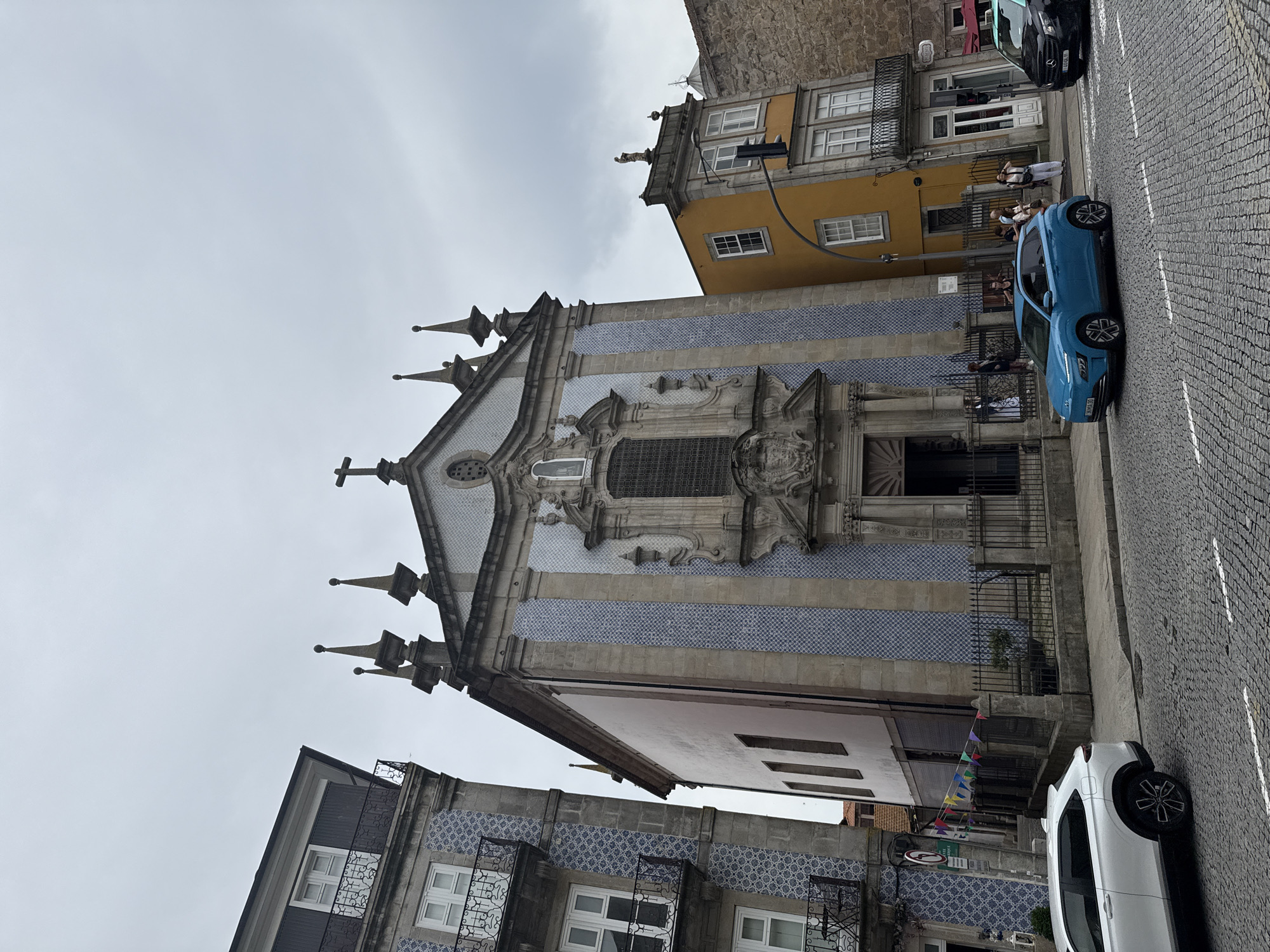 A blue car is parked on a cobblestone street in front of a historic building with a white and blue tiled facade. The building appears to be a church or a similar religious institution, with ornate architectural details and multiple spires. Adjacent to it is a yellow building with multiple windows and balconies. Several people are visible in the background, near the entrance of the yellow building, and on the street. A white car is parked further down the street, and a black car is partially visible behind it. Colorful flags are strung across the street in front of one of the buildings. The sky is overcast, suggesting a cloudy day. The scene is set in a European city, likely Porto, Portugal, given the architectural style and the mention of location context.