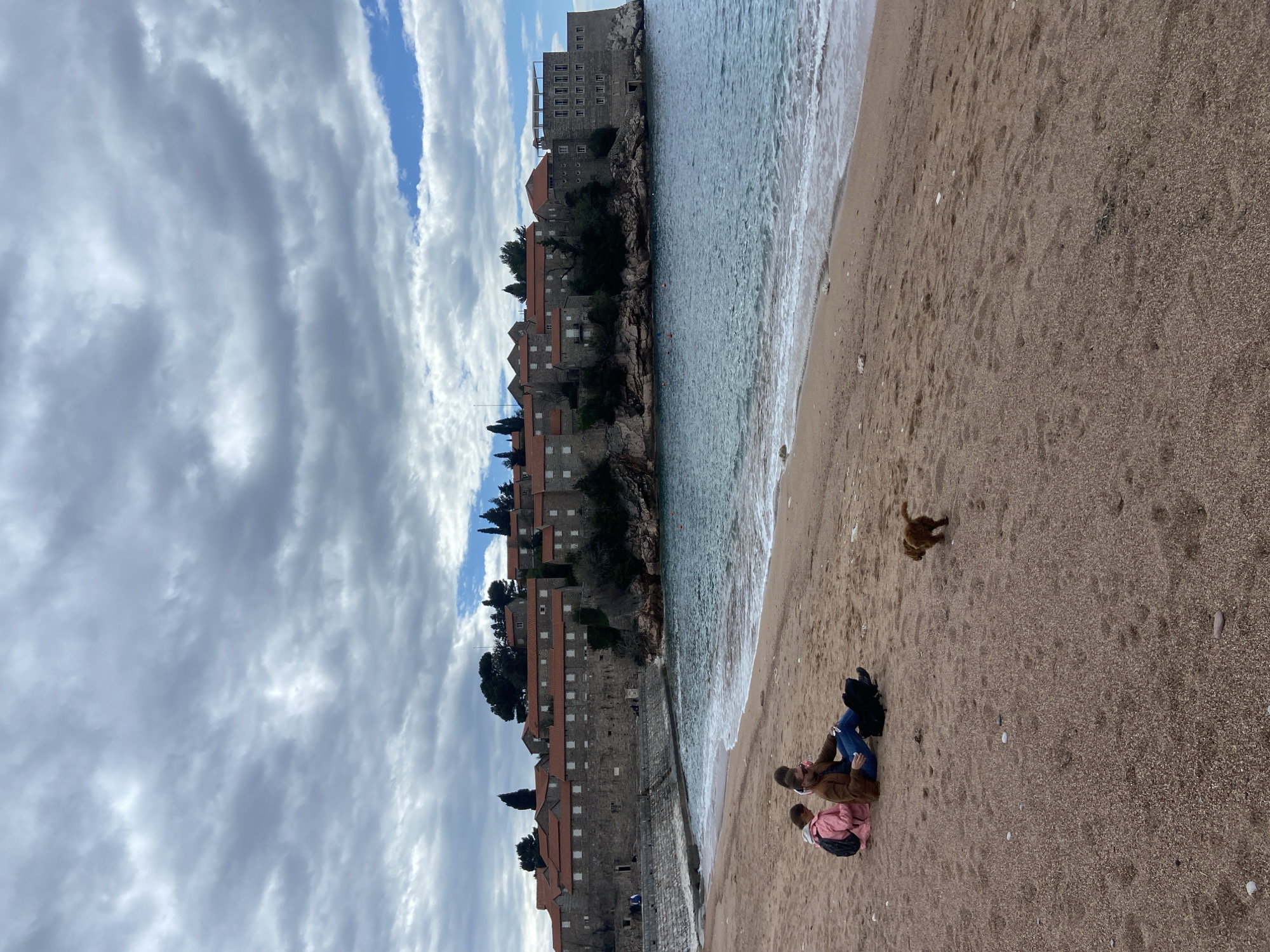 The image captures a beach scene in Sveti Stefan, Montenegro. The historic island town sits atop a rocky outcrop, with stone buildings featuring red-tiled roofs. The beach is sandy, bordering the clear turquoise water. Three people are sitting on the sand, engaged in conversation. A dog is running near them. The sky above is filled with streaky clouds, with patches of blue visible.