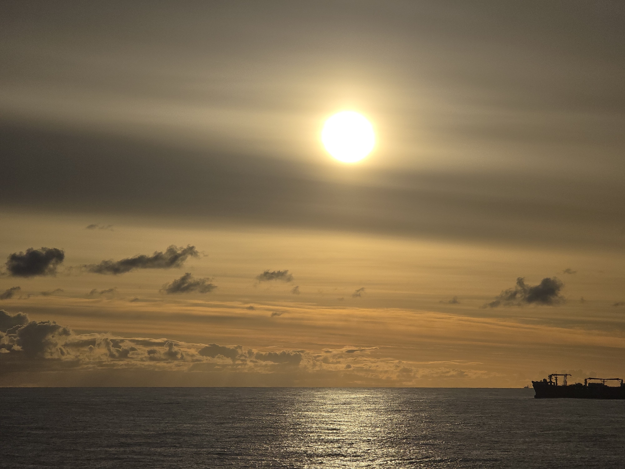 The image depicts a seascape with a prominent sun shining brightly in the sky. The sky is filled with clouds, some darker and some with an orange hue likely caused by the sun's rays. The ocean surface reflects the sunlight, creating a shimmering effect. A ship is visible on the horizon, silhouetted against the bright sky. The location is Unknown, Unknown. There are no people visible in the image.