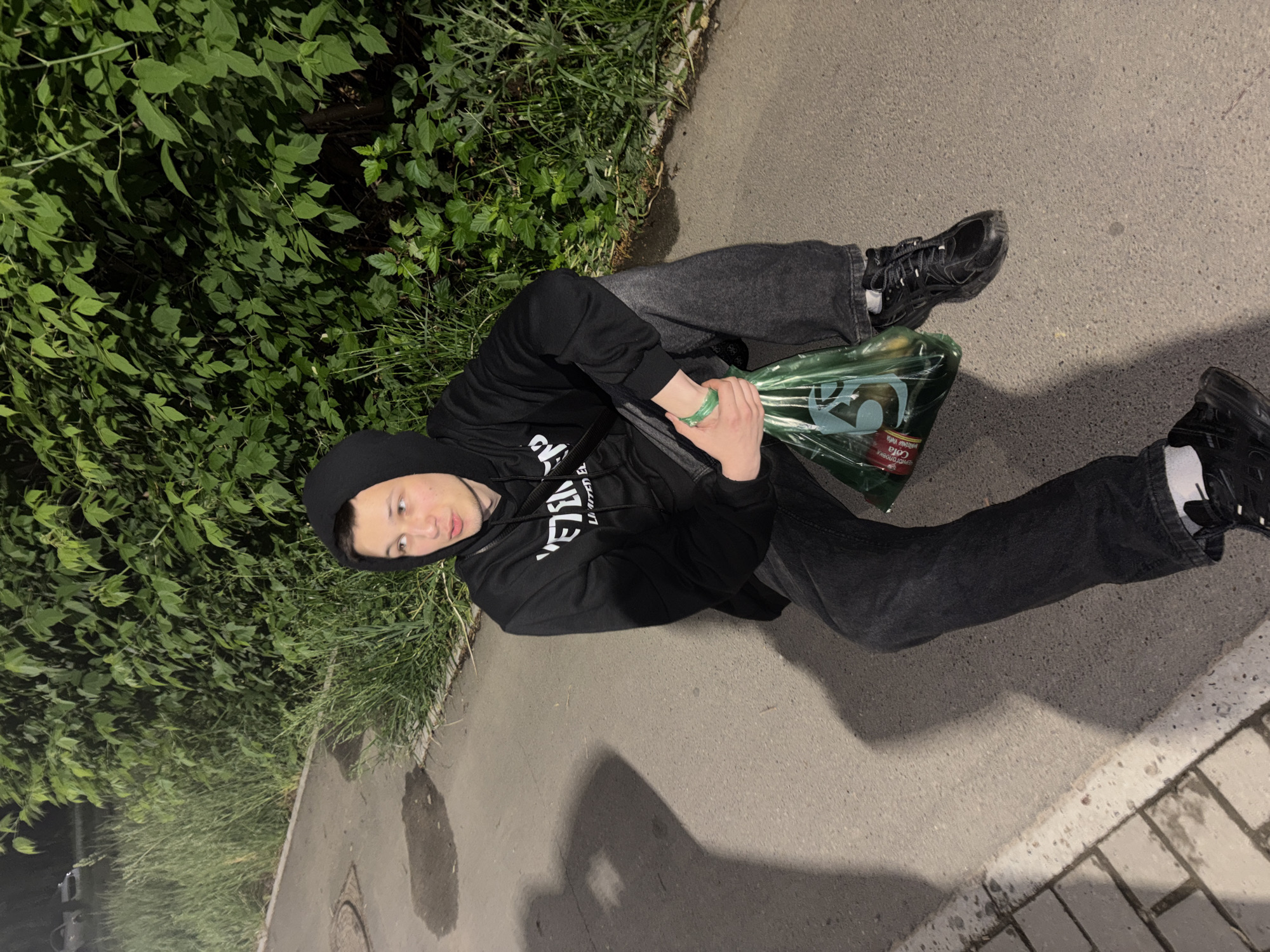 A young man wearing a black hoodie and dark jeans sits on a paved surface, likely a sidewalk or street, next to a patch of dense green foliage. He holds a large, clear plastic bag filled with various items, including what appears to be a can of cola. The man's left foot is visible with a white sock, while the other foot is black. The background features a gray concrete or asphalt surface. The image is taken at an angle, suggesting a high vantage point.
Based on the location, the surroundings do not offer many clues, but since the city is mentioned the picture may have been taken in Chaikovsky, Russia.