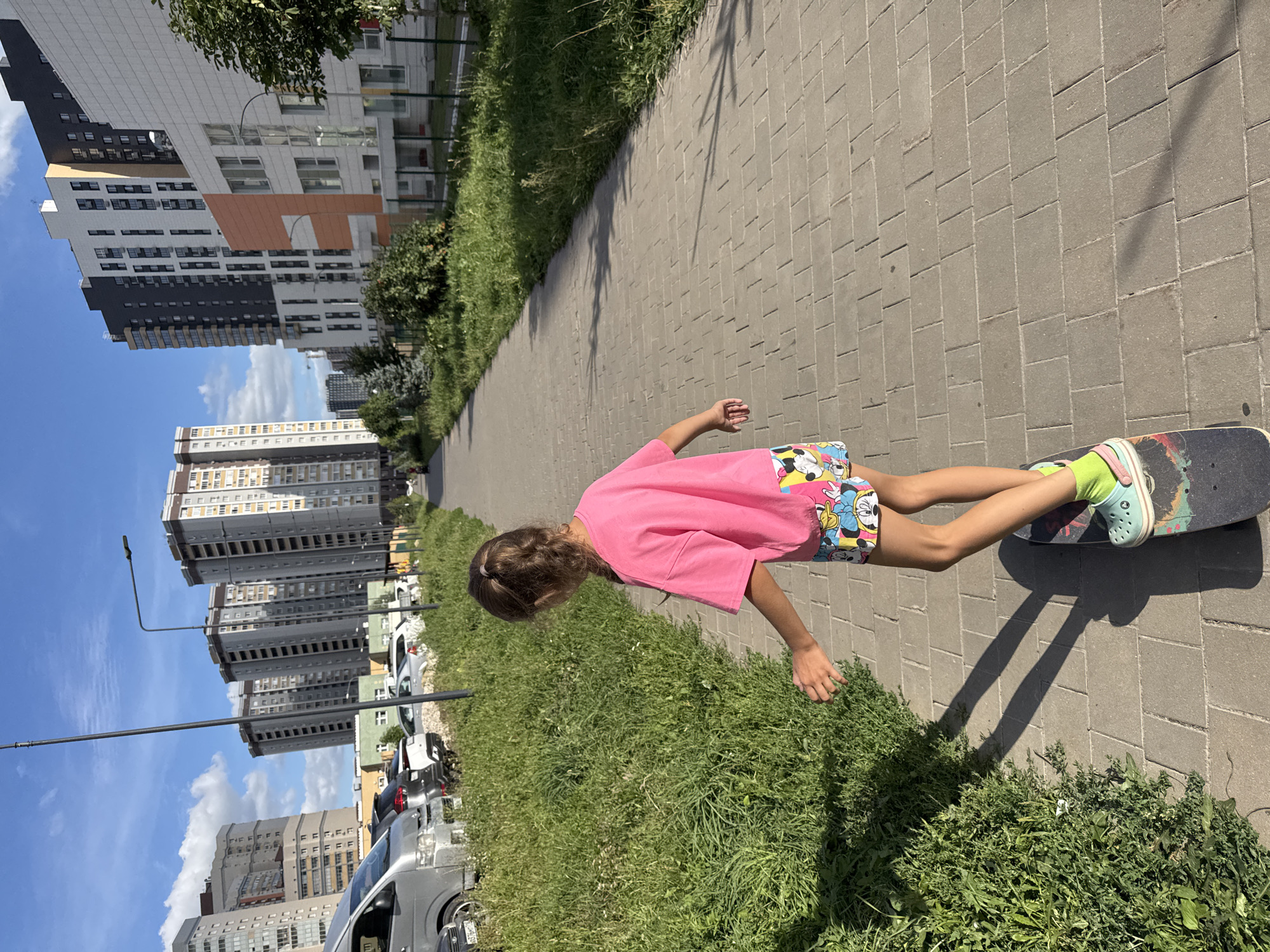A young girl with braided hair, wearing a bright pink t-shirt and colorful shorts with cartoon characters, is skateboarding on a paved sidewalk. She is facing away from the camera, her arms outstretched slightly for balance. Her shoes are light blue with lime green socks. The sidewalk is made of grey rectangular pavers, and it runs alongside a strip of lush green grass. In the background, there are several modern, multi-story apartment buildings with a mix of grey, white, and orange facades. The sky is blue with scattered white clouds, and the sun is casting shadows on the pavement. The scene appears to be a residential area in Kazan, Russia, possibly a newer development given the architecture.