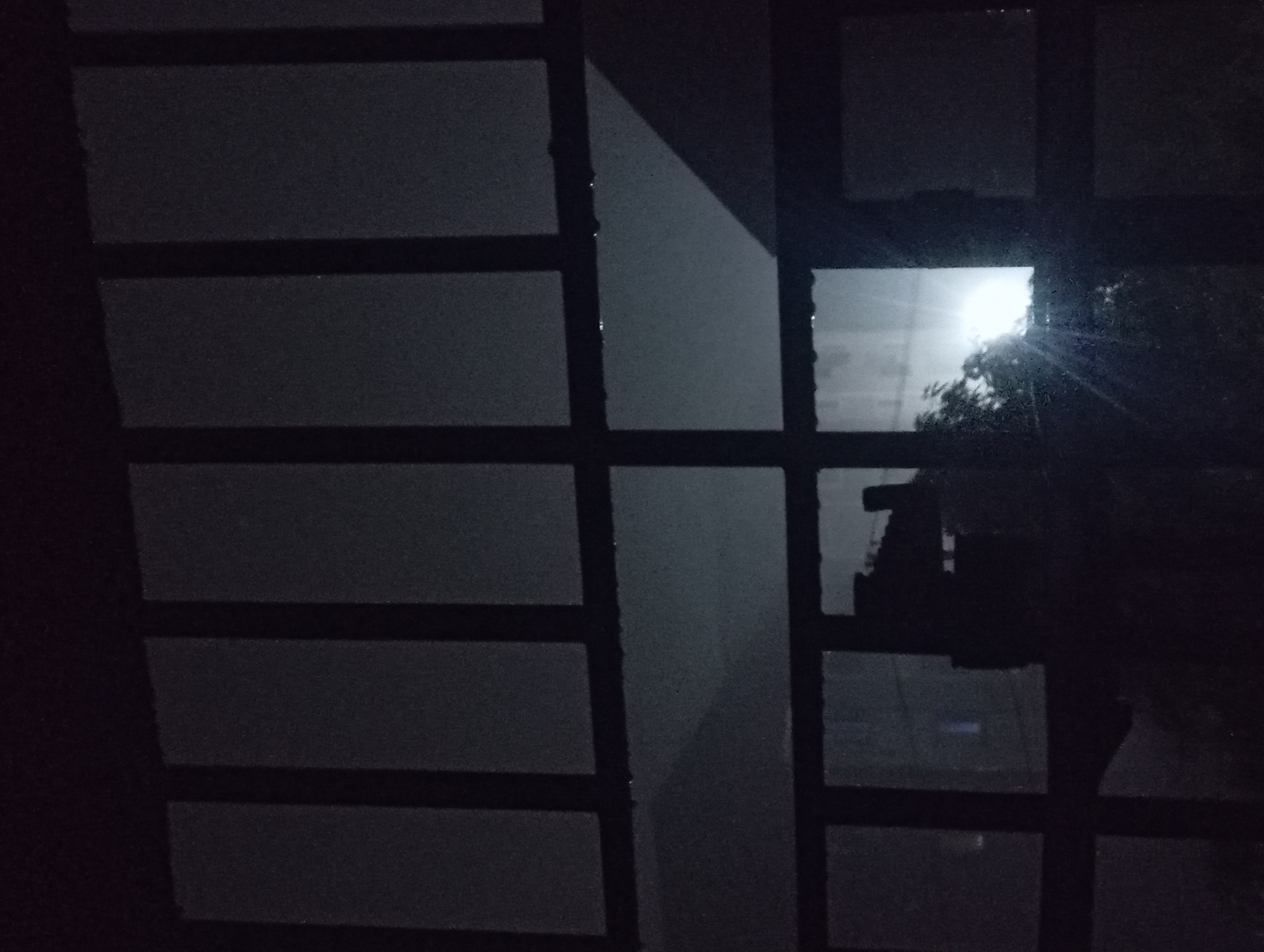 The image depicts a view from inside a building, looking out through a window with a grid-like frame. Outside, a light source, possibly the moon or a street lamp, shines through trees. The darkness and the grid-like window suggest a closed or semi-closed indoor space. There is no evidence to confirm the location as Nkpor, Nigeria, or to indicate any people, subjects, objects, actions, or interactions beyond the basic description provided.