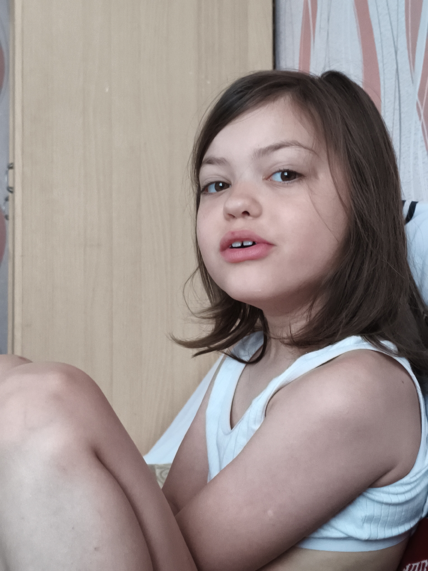 A young girl with brown hair is sitting on a chair. She is wearing a white tank top and is looking at the camera. The background is a wooden wall and a patterned wallpaper. The lighting is natural and the image is taken from a close-up perspective.