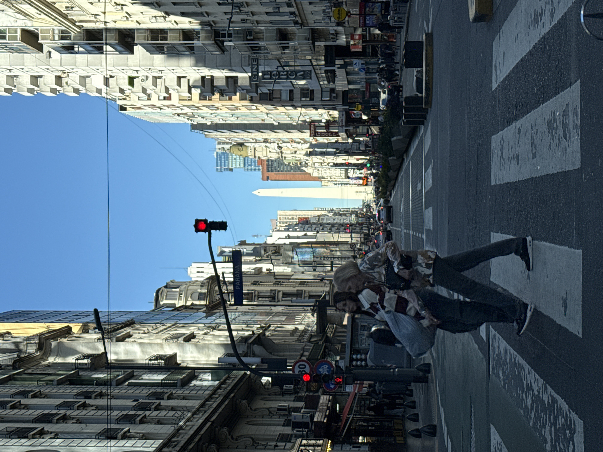 The image captures a vibrant street scene in Buenos Aires, Argentina, looking down a wide avenue flanked by tall city buildings under a clear blue sky.
In the foreground, on the right side of the street, several pedestrians are visible. One person, wearing a patterned jacket, dark pants, and sneakers, stands on a marked zebra crosswalk, suggesting they are waiting to cross. Red traffic lights are prominent in both the immediate foreground and further down the street, indicating that traffic may be stopped.
The avenue stretches into the distance, lined on both sides by numerous multi-story buildings, characteristic of a bustling urban environment. These buildings display a mix of architectural styles, with visible windows, balconies, and commercial signs. In the mid-ground, a very tall, white obelisk, the iconic Obelisco de Buenos Aires, stands prominently, confirming the location. Cars and other vehicles are visible further down the avenue.
The scene takes place during bright daylight, with strong, clear lighting and shadows indicating a sunny day. The sky is a cloudless, vivid blue.
Visible text includes: "PARANA" on a blue street sign, "CINE" and "LUCIA" on a building's facade, "Subte" on a yellow sign (indicating a subway entrance), "IDI," "PIZZERIA," and traffic signs displaying numbers like "30."