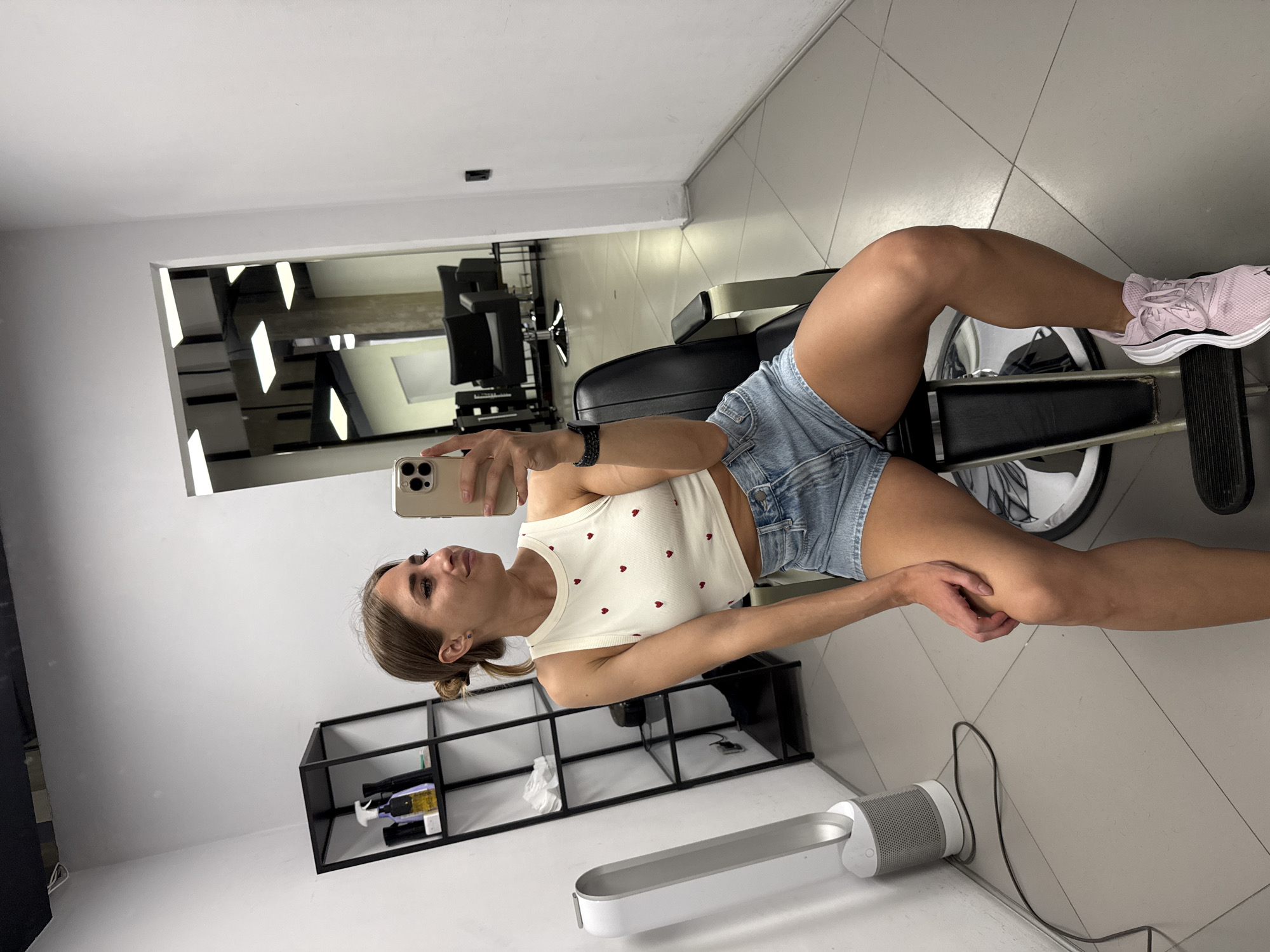 A young woman with brown hair pulled into a bun is taking a mirror selfie in what appears to be a barbershop or salon. She is wearing a white tank top with red hearts and denim shorts, and pink sneakers. She is sitting on a black barber chair. To her left, there is a tall, cylindrical white fan and a black shelving unit with bottles and other items. In the background, visible through an open doorway, are more barber chairs and mirrors. The walls are white, and the floor is tiled in a grey geometric pattern. The lighting in the room is bright and artificial.