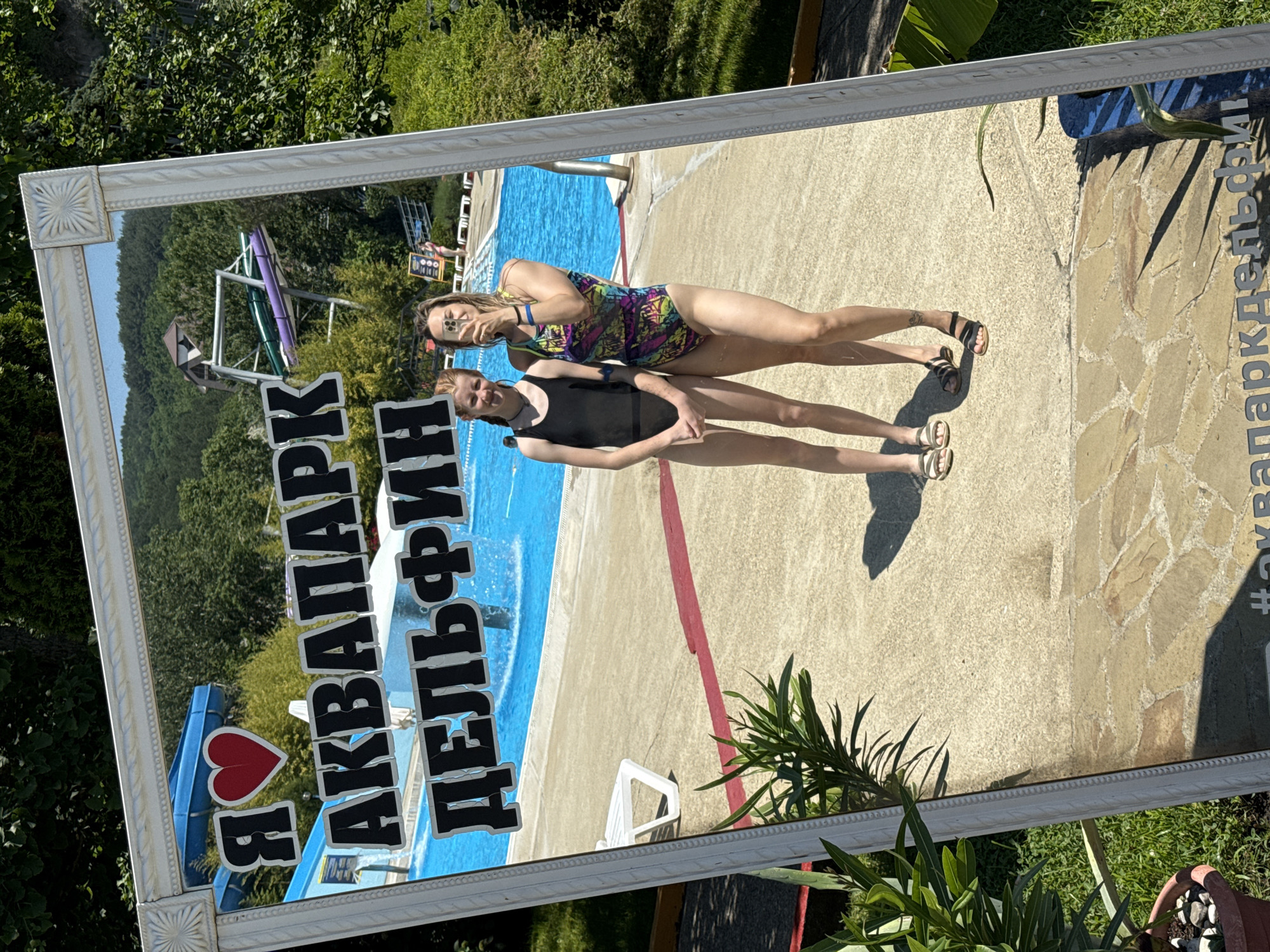 In the city of Nebug, Russia, at an aquapark, a framed mirror reflects two young women in swimsuits posing for a photo. The mirror is ornately decorated with a white frame. The reflection reveals a blue swimming pool with a water slide in the background. The text "Я ЛЮБЛЮ АКВАПАРК ДЕЛЬФИН" is prominently displayed on the mirror. The first woman, on the left, wears a black one-piece swimsuit. The second woman, on the right, wears a colorful, patterned one-piece swimsuit and holds a smartphone, taking the selfie. Both women are wearing sandals. The foreground shows a paved walkway and greenery, indicating an outdoor setting under a sunny sky. The overall scene captures a moment of leisure and fun at a summer water park.