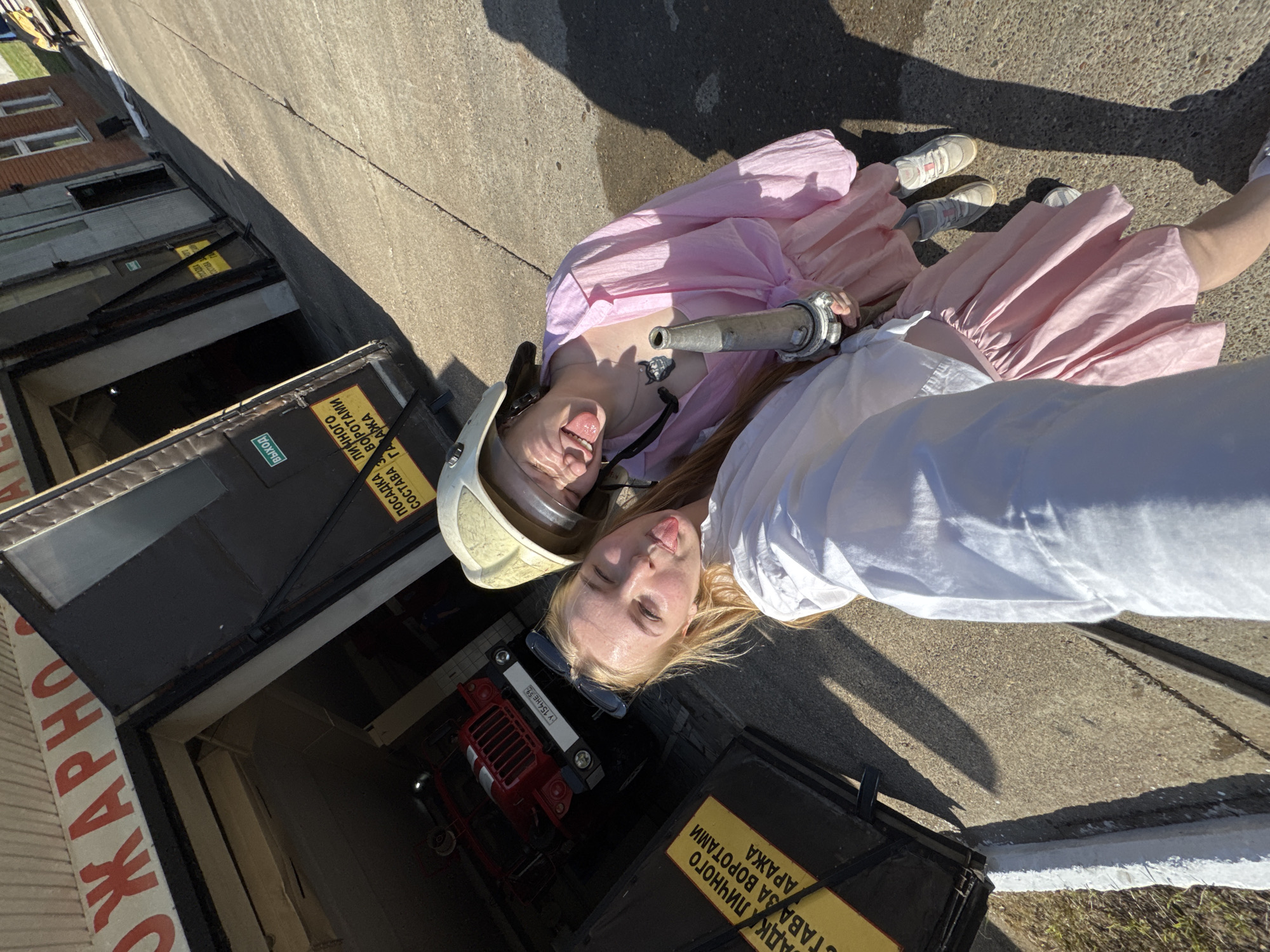 Two young women are lying on a concrete surface next to a building. One is wearing a pink dress and a fire helmet. The other is wearing a white shirt. They are both sticking out their tongues. Between them is a metal fire hose nozzle. Behind them, there is a building with garage doors. One garage contains a small red vehicle. The garage doors have signs in Cyrillic script on them. The text contains the word “ГАРАЖА” (GARAGE). One license plate says "Y 154 HE 59". The location may be Rusalevka, Russia, based on the Cyrillic script.