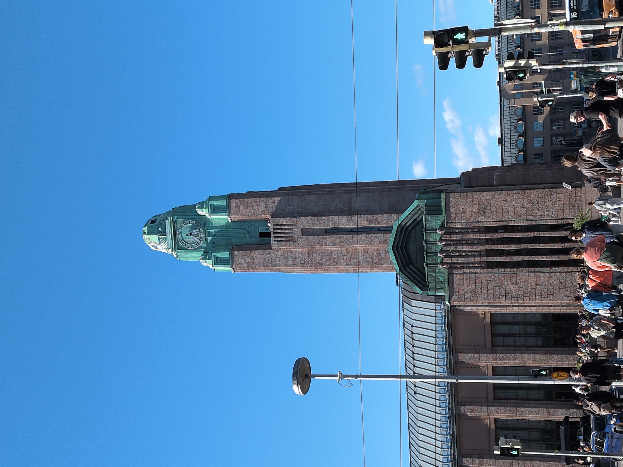 This image captures the Helsinki Central Railway Station in Helsinki, Finland, under a clear blue sky with scattered clouds. The iconic clock tower, made of red granite and adorned with green copper detailing, dominates the center of the frame. The clock face is visible, showing approximately 11:30. The station building features a long, arched roof with a metal surface, and the facade is constructed from the same red granite as the tower.
In the foreground, a diverse group of pedestrians is walking on the street. They are of various ages and ethnicities, dressed in casual summer clothing. Some are walking together, while others are alone. Several vehicles are also present, including a blue car and a yellow and orange bus with the number "16" displayed on its side. Traffic lights are visible, with one showing a green pedestrian signal. A street lamp with a circular reflector stands to the left of the tower. The overall scene depicts a busy urban environment, typical of a city center on a sunny day.