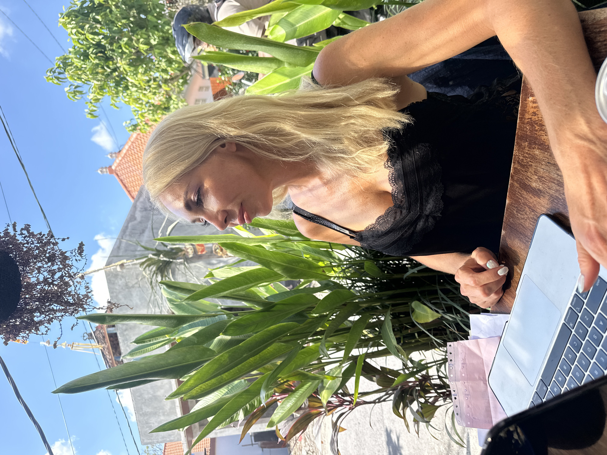A blonde woman sits at a wooden table outdoors, facing right and looking away from the camera. She wears a black camisole with lace trim. Her hands are on the table near a laptop, one resting on the table and the other touching the laptop keyboard. In the background, lush green foliage and tropical plants are visible, along with buildings and a clear blue sky with clouds. A motorcycle rider is partially visible on the right side of the image.