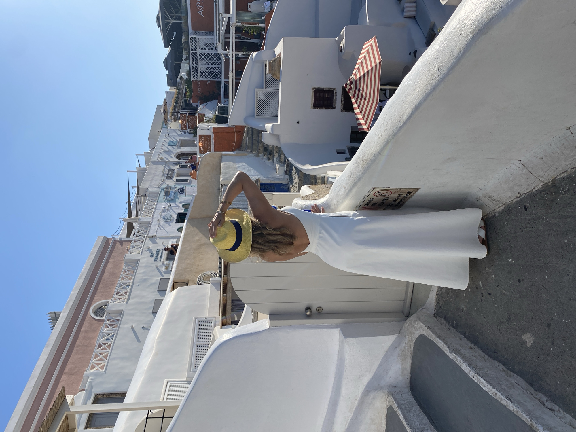 A woman wearing a white dress and a yellow hat stands on a white balcony in Oia, Greece, facing away from the camera and looking at the view. The architecture is typical of Oia, with whitewashed buildings, blue accents, and stairs winding through the town. The sky is clear and blue, indicating a sunny day. A striped umbrella is visible in the background.