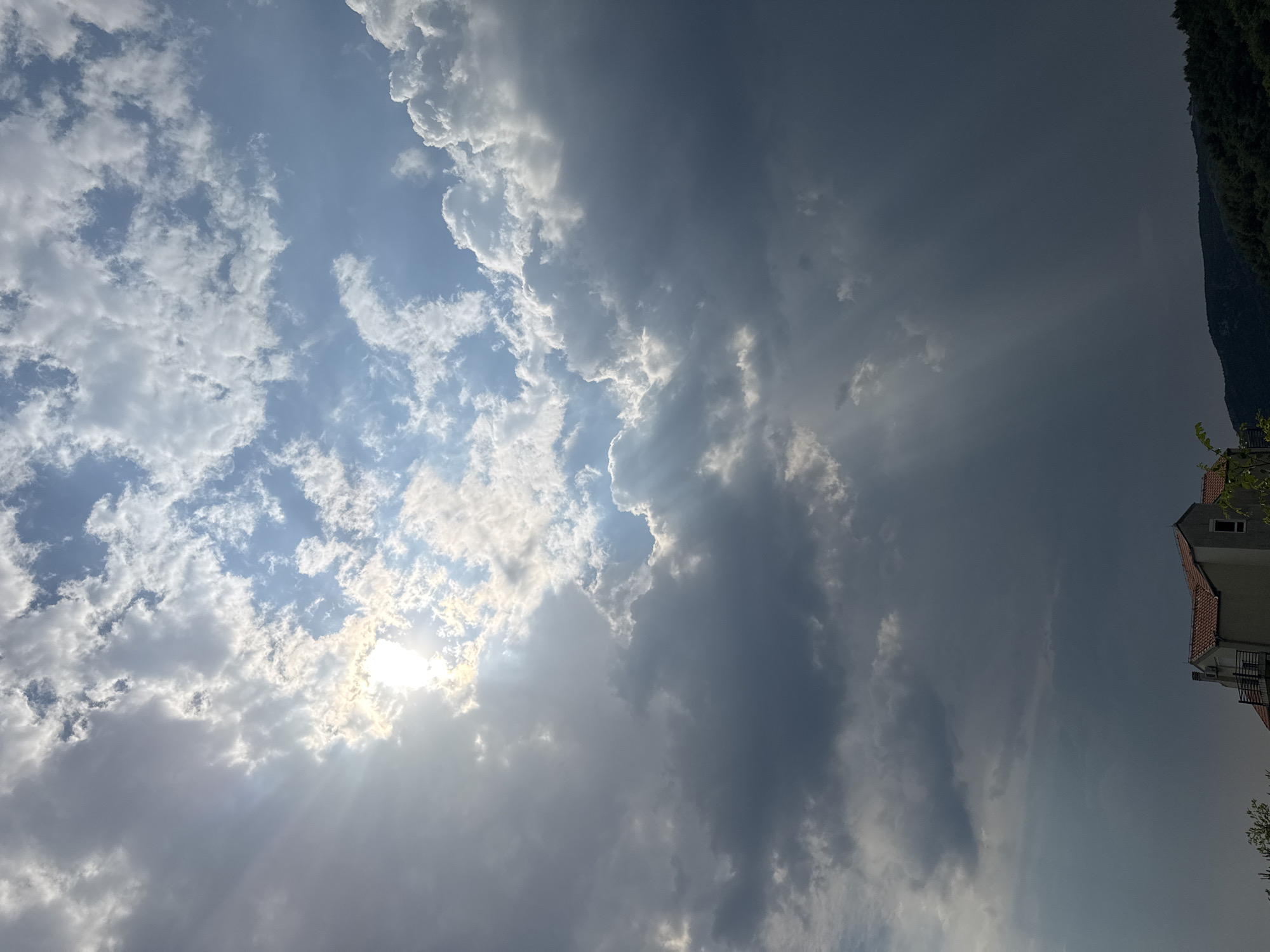 This media file is an image capturing a dramatic sky over Rafailovići, Montenegro. The primary subject is the sky, filled with a dynamic interplay of light and shadow cast by a mix of bright white cumulus clouds and darker, more ominous storm clouds. Sunlight pierces through the clouds, creating visible sunbeams that add a sense of divine light to the scene. The overall weather appears to be changing, with a blend of clearing skies and approaching rain suggested by the darker cloud formations.
In the upper right corner, a portion of a building with a red-tiled roof and a dark roofline is visible, along with lush green foliage. This building, along with the presence of a steep incline in the far right, suggests a coastal or hillside location, consistent with Rafailovići, Montenegro.
The time of day is likely late afternoon or early evening, given the angle and quality of the sunlight, which is casting long rays. The emotional tone of the image is one of awe and perhaps a touch of foreboding, due to the contrast between the bright, illuminated parts of the sky and the darker, heavier clouds. No people or specific objects are prominently featured, as the focus is on the natural spectacle of the sky. There is no visible text in the image.