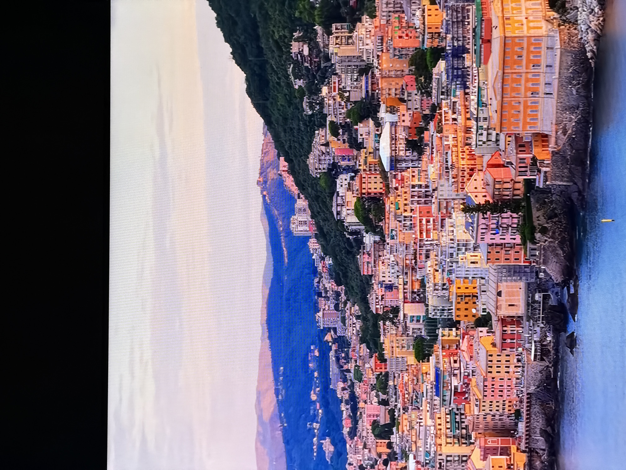 This image depicts a coastal city with colorful buildings cascading down a hillside towards the water. The architecture is dense, with many multi-story structures painted in shades of orange, pink, yellow, and red. Some buildings appear to be apartments or hotels, while others might be residences. The city is nestled against a steep, green hillside covered in trees, with more distant, blue-hued mountains forming the backdrop. The sky is a pale, hazy blue with hints of pink, suggesting either sunrise or sunset. The water in the foreground is a deep blue, with small rocks visible near the shore. There are no people or vehicles clearly visible in the image. The scene conveys a sense of a vibrant, densely populated European coastal town. This description is not consistent with the city of Chirchik, Uzbekistan, which is an inland city.