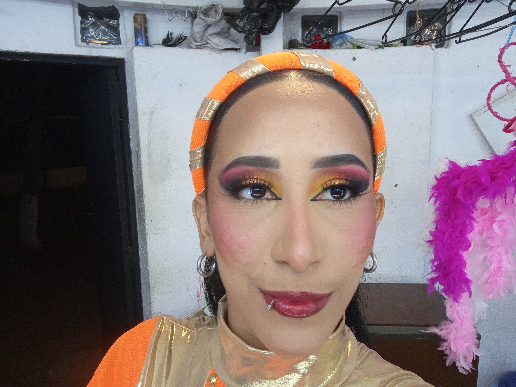 A close-up shot captures a person with dark hair and heavy stage makeup. They wear an orange and gold headband and gold top with orange shoulders. The makeup features bold eyeshadow in shades of red, orange, and gold, as well as heavy eyeliner and false eyelashes. They also wear bright red lipstick and blush. A lip piercing is visible on the lower lip.
The background is a white, textured wall with multiple small alcoves, some containing items. A dark doorway is visible on the left. A feather boa in shades of pink and purple hangs on the right.
Without further information, it is impossible to confirm the location as Porlamar, Venezuela.