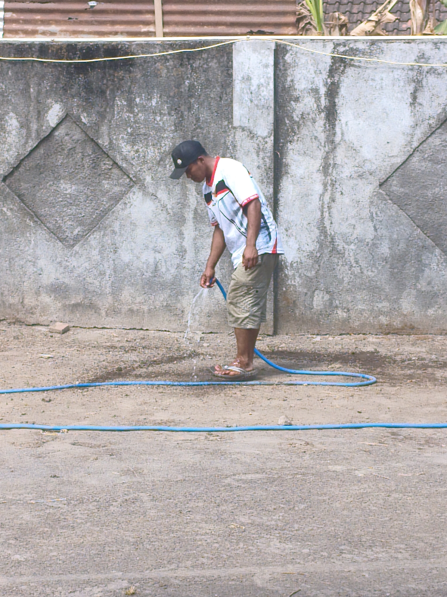 A man wearing a black baseball cap, a white shirt with red and black accents, and khaki shorts, is watering the ground with a blue hose. He is standing in front of a gray, textured concrete wall with a diamond-shaped pattern. The ground is dirt and gravel, with a blue hose laid out on it. In the background, a corrugated metal roof and some tropical foliage are visible. The scene appears to be outdoors in a dry, possibly urban or semi-urban environment, potentially located in Berbah, Indonesia, given the architectural and environmental cues.