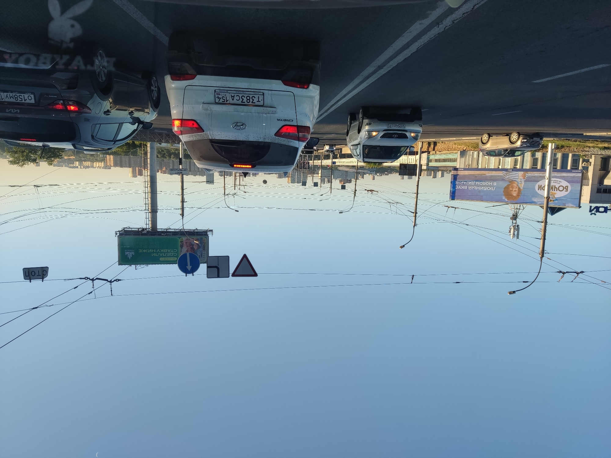 This media file captures a view of a city street in Novosibirsk, Russia, likely during the daytime due to the bright sky. The image is oriented upside down, showing cars and billboards from a perspective below street level.
In the foreground, traffic signals and overhead power lines are prominent. Several cars are visible on the road, including a white Hyundai Creta with a Russian license plate "T383CA 154" and a dark-colored Kia with a license plate "0158MY 1". The road appears to be a multi-lane highway with clear lane markings.
Further down the street, large billboards are erected. One billboard, with the text "БОЧКОРИ" and "ЛЮБИМЫЙ ВКУС в новом дИЗАЙНЕ" (Bochkori, Favorite Taste in a New Design), features a smiling child. Another billboard displays the text "СТАВКУ НИЖЕ" (Lower the Rate). The city skyline with numerous buildings is visible in the background, suggesting an urban environment.
A "STOP" sign in Russian, "СТОП", is also visible. The overall impression is that of a bustling city street with ongoing traffic and commercial advertising. The clear blue sky indicates fair weather.