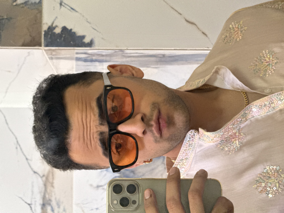 A male individual is depicted in a mirror selfie taken indoors in Malasar, India. The subject, a young man with dark, styled hair, is seen from the chest up, facing the camera's reflection. He wears black-framed sunglasses with prominent orange-tinted lenses. His left eyebrow is raised, and his lips are slightly pursed. A small gold-colored hoop earring is visible on his left ear. He is dressed in a light-colored, possibly off-white or beige, traditional Indian kurta featuring intricate, multi-colored embroidery and sequin work along the mandarin collar and front placket, extending onto the chest. A gold-colored cuban link chain is visible around his neck beneath the open collar. In his left hand, which is visible in the lower-left foreground, he holds a light-colored smartphone with a visible triple-lens camera array, consistent with an iPhone model, used to capture the image. The background consists of tiled walls, featuring white marble-patterned tiles on the left and a combination of white, grey, and possibly blue marbled tiles on the right, separated by a vertical grout line, suggesting an interior location such as a bathroom or dressing area.