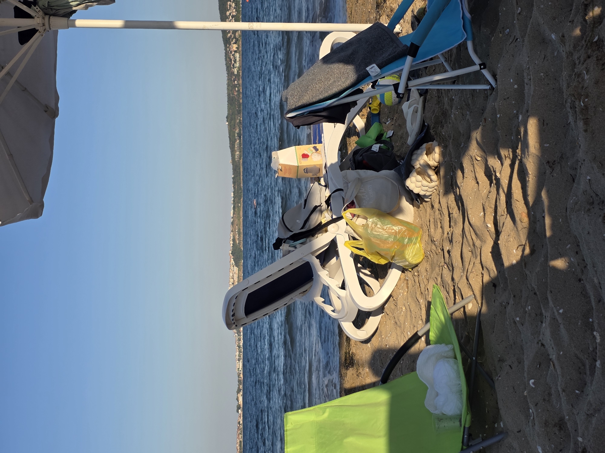 A sun-drenched beach scene in Chernomorets, Bulgaria, shows the aftermath of a day at the seaside. Two beach chairs are set up on the sand, one lime green and the other blue. The green chair in the foreground is occupied by a pile of white towels and two plastic cups. To its right, a white plastic lounge chair is laden with various items, including a yellow plastic bag, a beige canvas tote bag, a small cardboard box, and a gray blanket. A large white beach umbrella provides shade over part of the scene. In the background, the calm blue sea stretches towards a distant shoreline dotted with buildings and lush green hills. The sky is a clear, brilliant blue. The sandy beach is scattered with small seashells and debris. The overall impression is one of a deserted beach after a day of activity.
