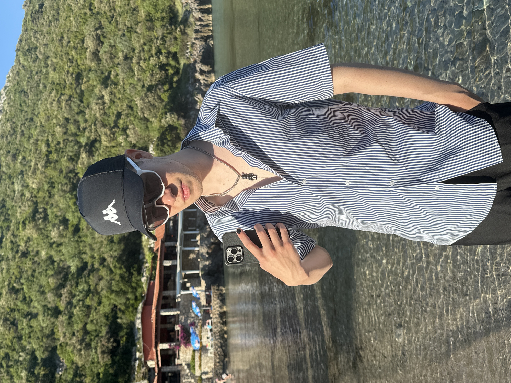 In Lastva Grbaljska, Montenegro, a young male-presenting individual stands in clear, shallow water near a shore under bright sunlight. He is wearing a black baseball cap with a white Kappa logo, white-framed sunglasses, and a short-sleeved shirt with vertical navy blue and white stripes, unbuttoned at the top to reveal a silver chain with a black bear-shaped pendant. His left hand holds a dark smartphone, identified by its triple camera array, vertically in front of him; his fingernails are painted black. His right hand is partially inserted into a pocket of his dark trousers. The background features a large hillside densely covered with green vegetation. To the left, a complex of stone buildings with reddish roofs, possibly a resort or restaurant, is visible near the water, with blue umbrellas and lounge chairs on a paved area. The water in the foreground is clear, showing ripples and the seabed.