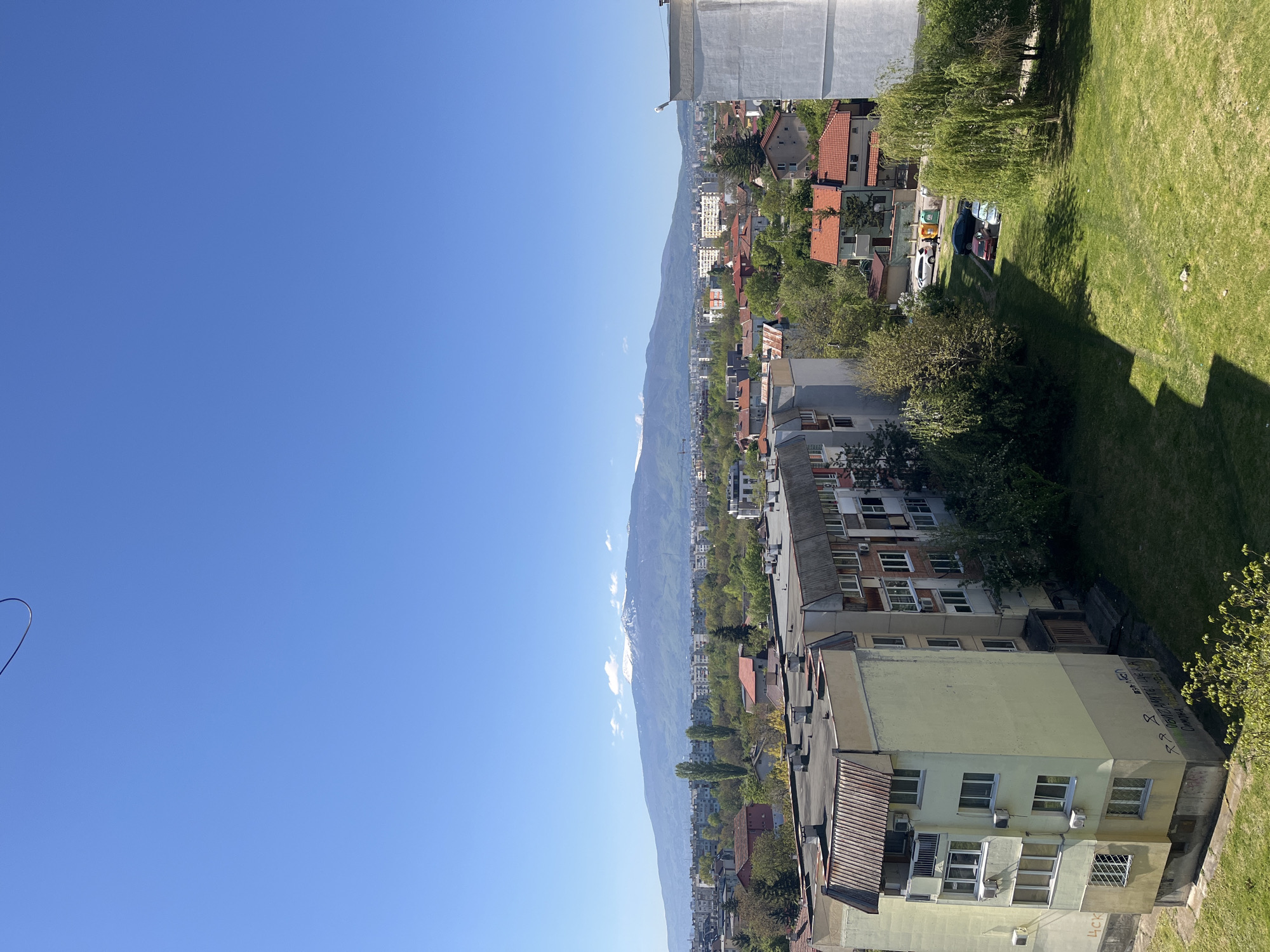 A medium-level, outdoor shot depicts a cityscape under a clear blue sky. The buildings are of varying heights and styles, interspersed with trees and green spaces. In the distance, a mountain range can be seen, indicating the presence of a natural landscape nearby. The location is likely Sofia, Bulgaria, given the architectural style and mountainous backdrop common to the city. The scene appears to be taken from an elevated position, possibly a high-rise building or hill, offering a panoramic view of the urban landscape. The overall atmosphere is bright and sunny, suggesting a pleasant day in the city.
