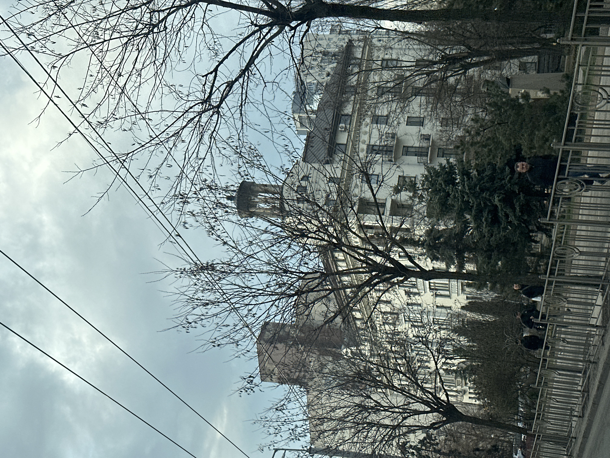 A street scene in Kyiv, Ukraine, is captured on an overcast day. A row of tall, white apartment buildings with multiple windows dominates the background, interspersed with bare trees. One building features a distinctive tower. Power lines stretch across the upper left of the frame. A metal fence runs along the right side of the image, partially obscuring a group of people standing behind it. The ground is paved, suggesting a sidewalk or street.