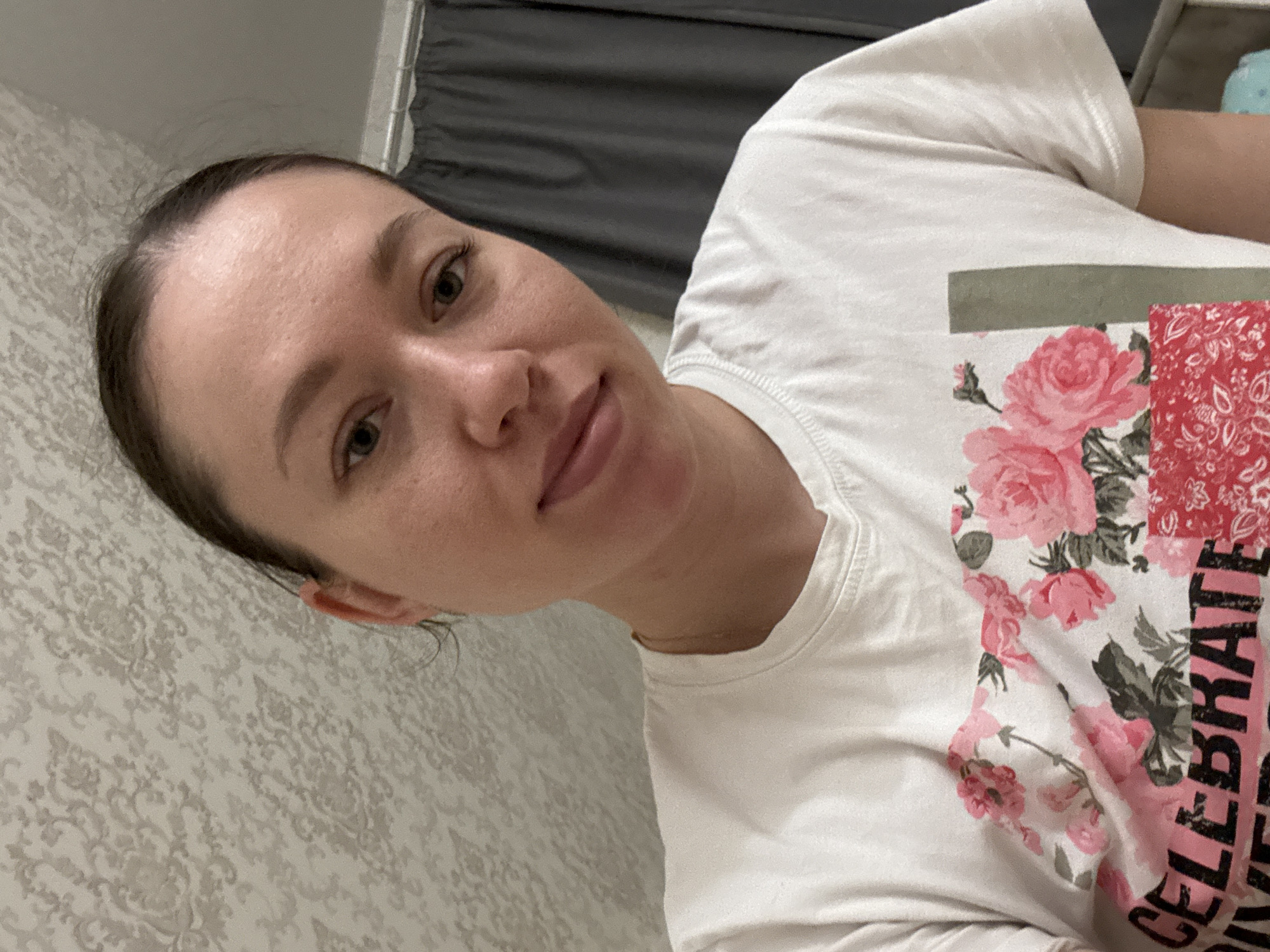 The image depicts a woman with fair skin and dark hair pulled back from her face. She is wearing a white t-shirt with a floral and paisley design. The word "CELEBRATE" is partially visible on the shirt. The woman is indoors, in front of a patterned wall and what appears to be a window with closed blinds. The location in the city of Ферма, Russia, cannot be determined from the image.