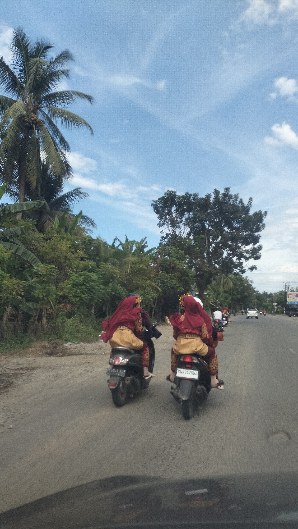 The image shows two individuals riding on motorcycles down a paved road in Glee Gogo, Indonesia. Both individuals are wearing traditional red and gold attire, including head coverings. The motorcycle on the left has a license plate that reads "PL 5913 VUR", and the motorcycle on the right has a license plate that reads "PL 4352 PRZ". Lush green foliage and palm trees line the side of the road. The sky is blue with scattered white clouds. In the distance, there are other vehicles and some buildings, suggesting a rural or semi-urban setting. The perspective suggests the photo was taken from another moving vehicle.