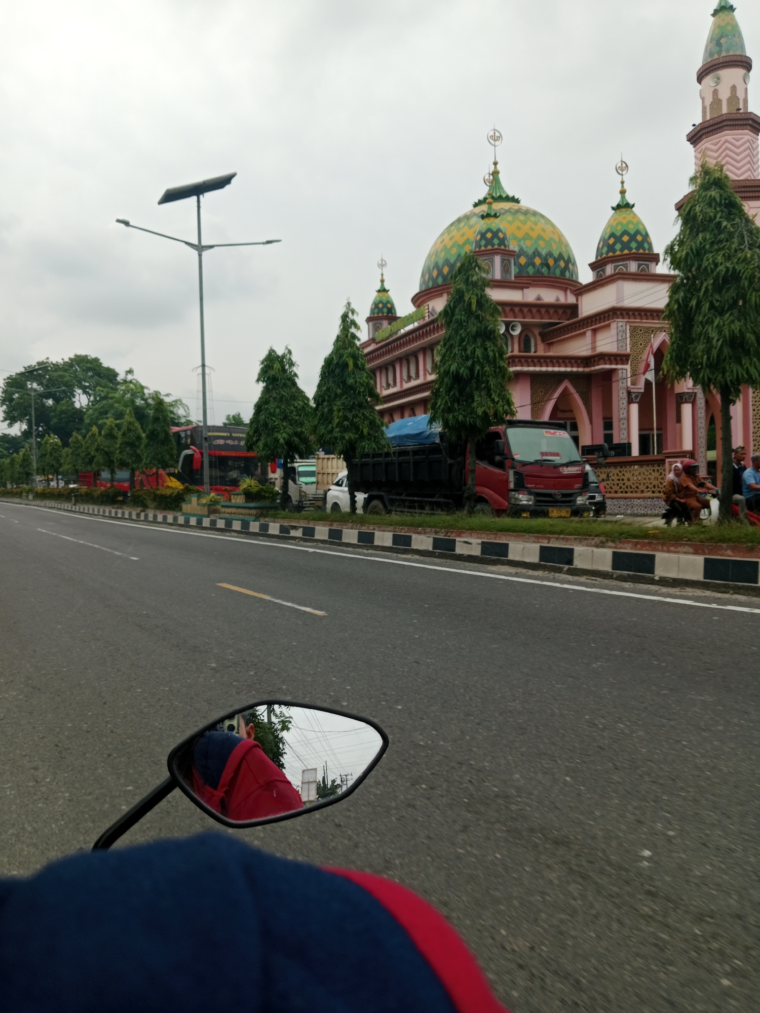 The image shows a street scene in the City of Binjai, Indonesia, with a prominent mosque in the background. The mosque has a distinctive pink facade and a series of green and yellow domes, with a tall, slender minaret. Trees line the street in front of the mosque, and a decorative fence separates the road from the mosque grounds.
On the street, several vehicles are visible, including a red truck with a blue tarp on its bed, a white truck, and what appears to be a bus. The road is paved and has lane markings. In the foreground, the reflection of a person wearing a red and blue jacket can be seen in a motorcycle's side mirror. The sky is overcast. A solar-powered street lamp is also visible on the left side of the image. People are seen on a motorcycle to the right of the red truck, some of whom are wearing headscarves.