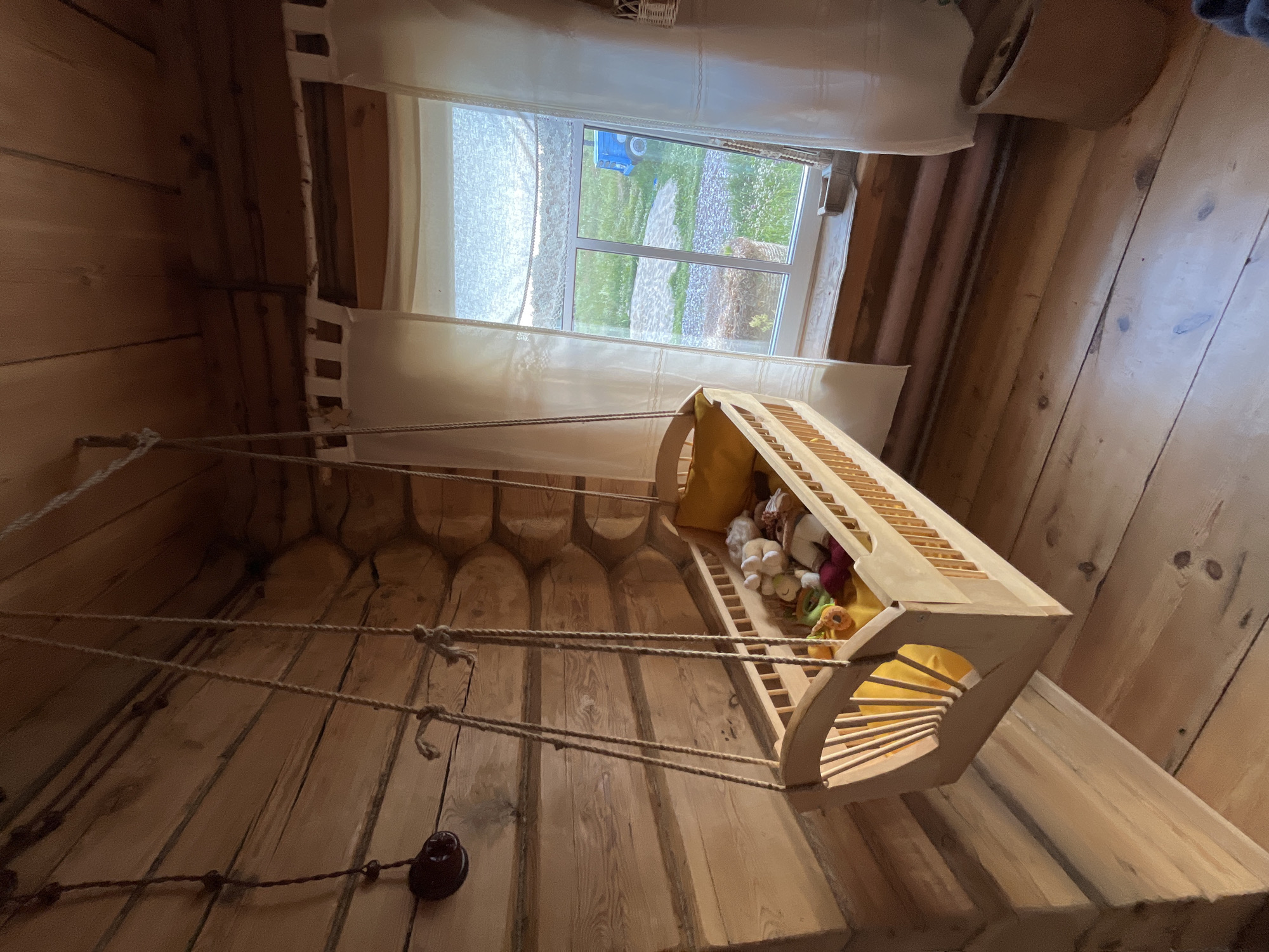 This image shows an interior scene of a room with wooden walls and floors. Suspended from the ceiling by ropes is a wooden cradle filled with stuffed animals. The cradle has a yellow interior lining and appears to be a child's toy or bed. Through a window in the background, a green, grassy area with some gravel paths and what appears to be a blue toy truck is visible, suggesting an outdoor environment. The window is covered by sheer white curtains. The lighting suggests it might be daytime. The room has a rustic aesthetic, with the wooden logs forming the walls being particularly prominent. There is a dark, circular object attached to the wall near the bottom left, which could be a light fixture or a similar mounting. The perspective of the photo is from a low angle, looking upwards towards the cradle and the window. The overall impression is one of a cozy, perhaps child-friendly, space within a wooden structure.