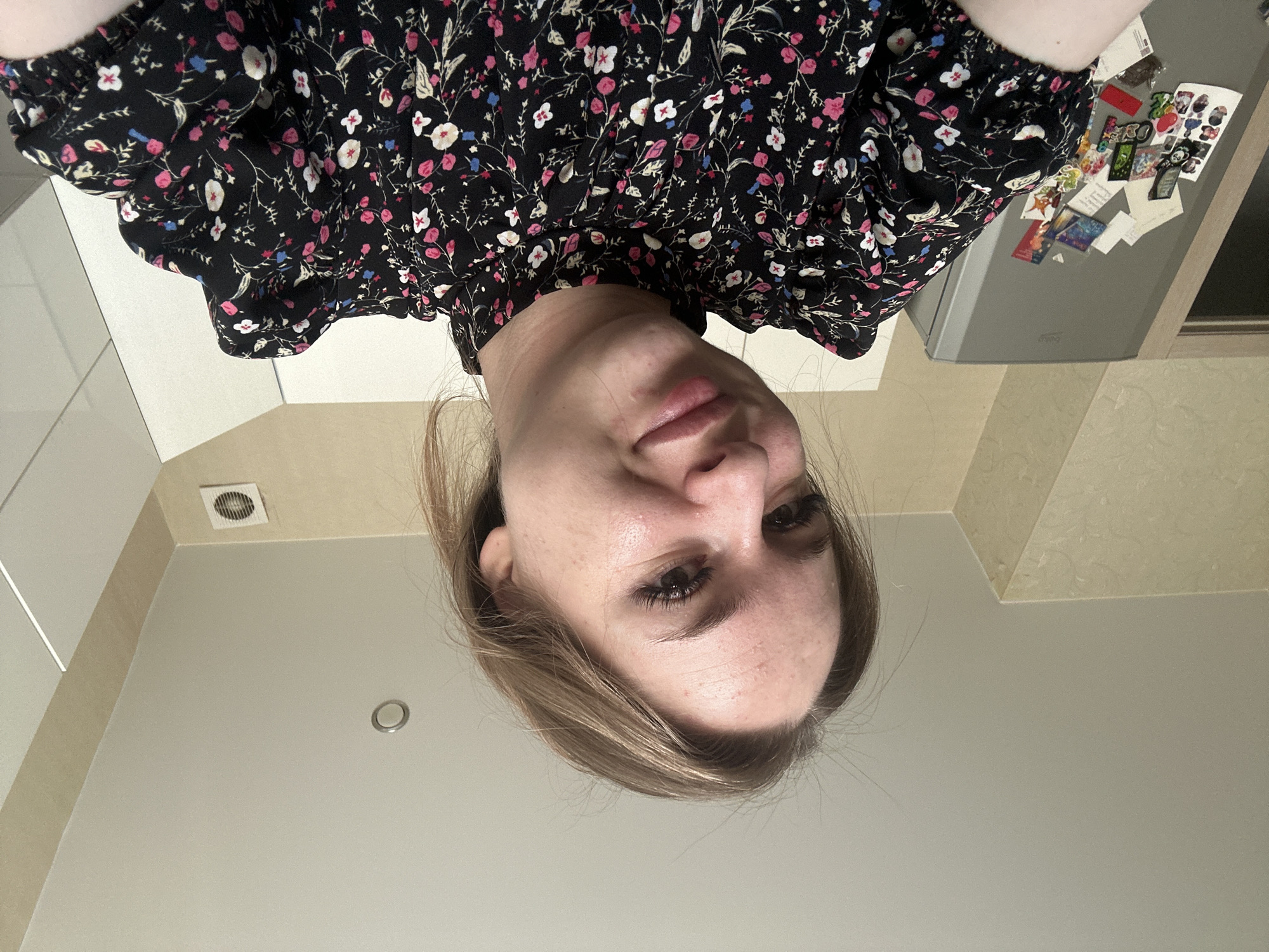 A low-angle selfie captures a Caucasian female, estimated to be in her 20s or 30s, smiling directly at the camera. She has light brown hair, dark eyes with prominent eyelashes, and visible skin texture on her forehead and cheeks. Her lips are slightly parted in a smile, showing some redness on the lower lip. She is wearing a black, high-necked blouse with puffed sleeves, adorned with a pattern of small pink, white, and blue flowers with green stems.
The scene is indoors, located in the city of Unknown, Unknown. The background features a light beige/off-white ceiling with a silver-colored recessed light fixture visible on the right side. The walls include a textured beige wallpaper on the left and a smooth white surface, possibly cabinetry, on the right. A square white ventilation grate is present on the lower right wall. To the left of the subject, a gray, two-door refrigerator is visible, heavily decorated with an assortment of magnets, stickers, and papers, including photographs and what appear to be cartoon or anime characters. A light-colored doorframe is partially visible on the far left.