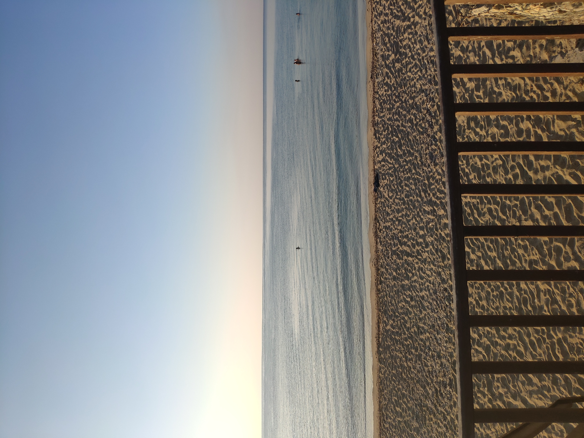 A tranquil beach scene unfolds under a clear, pale blue sky. In the foreground, a wooden railing made of vertical posts and horizontal beams separates the viewer from a sandy beach. The sand is imprinted with numerous small footprints and patterns, indicating recent activity. Beyond the beach, the calm, blue sea stretches out to a distant horizon, where it meets the sky in a soft, hazy line. A few people are visible in the water, appearing as small figures in the distance. The overall atmosphere is serene and peaceful, suggesting an early morning or late afternoon setting. The location is identified as Trikomo, Northern Cyprus / Cyprus.
