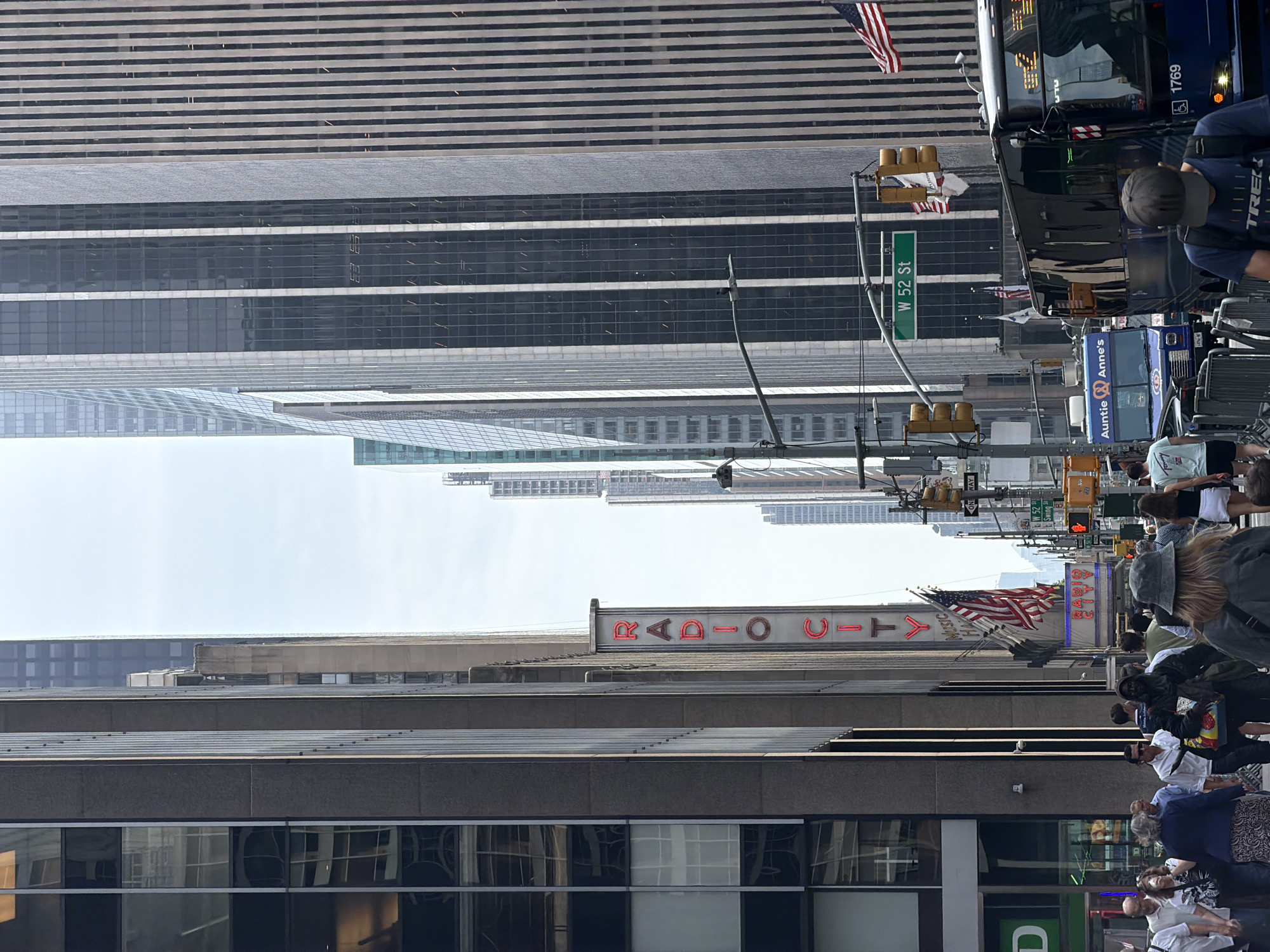 The image depicts a street scene in New York City, specifically along Sixth Avenue in Midtown Manhattan, looking north. The iconic Radio City Music Hall marquee and vertical sign are prominently visible, indicating the location. A diverse crowd of pedestrians is walking on the sidewalk, some appearing to be tourists. Several tall, modern office buildings line the street, with their glass and concrete facades reflecting the sky. Traffic signals and street signs, including one for "W 52 St," are present. A blue tour bus, labeled "Auntie Anne's," and a dark-colored bus with the number "1769" are stopped in traffic. American flags are displayed on poles. The weather appears overcast with some daylight.