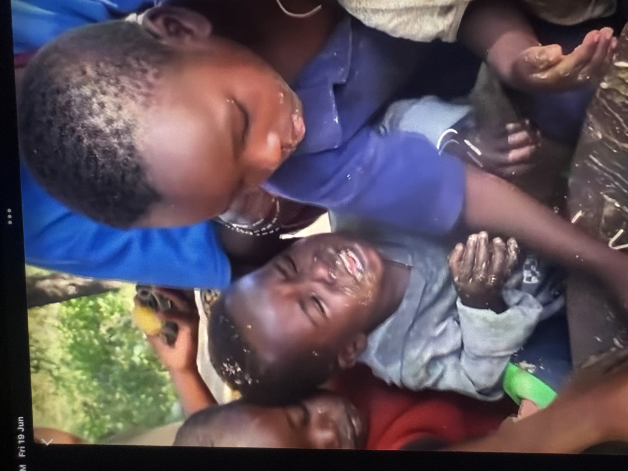 The image depicts a close-up of several young Black children in what appears to be an outdoor setting with greenery in the background. One child in the foreground, wearing a gray shirt, is crying and has food or dirt smeared on their face and hair. Another child, wearing a blue shirt, is leaning over the crying child. Other children are partially visible in the frame. The scene appears to be candid and shows the children in a raw, unposed moment. There is no information in the image that allows for the identification of the city of Abuja, Nigeria.