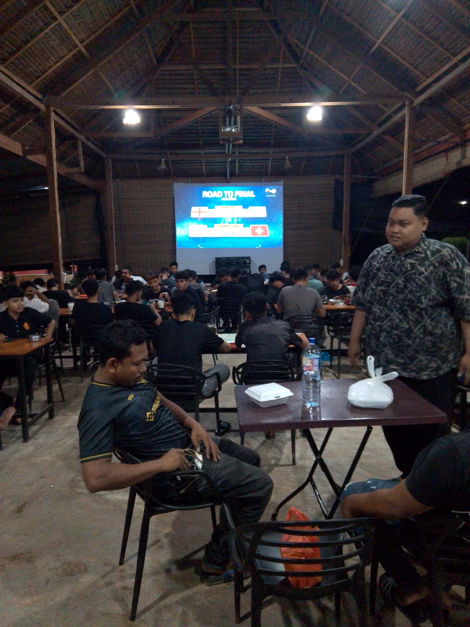 The image shows an indoor gathering of people in what appears to be an open-air restaurant or a large common area with a thatched roof. A projection screen displays "ROAD TO FINAL" along with country flags and text. Several individuals are seated at tables, some watching the screen, others engaged in conversation. In the foreground, a man in a dark, patterned shirt stands near a table with a bottle of water and a white plastic bag. Another man in a dark t-shirt is seated at a table on the left, holding a phone. The environment is dimly lit, with overhead lights and the glow from the screen illuminating the scene. The overall atmosphere suggests a relaxed social event, possibly related to a sports match or competition.