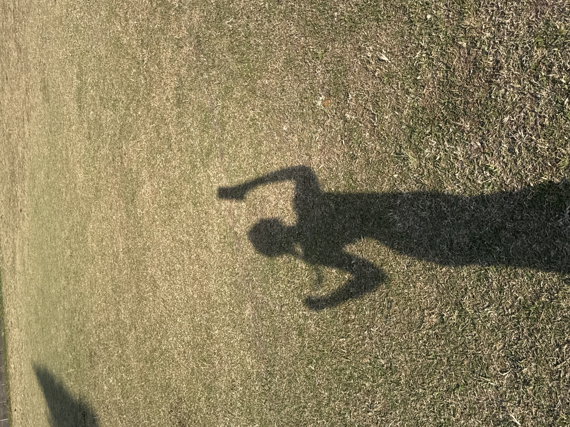 A shadow of a person, possibly a woman given the silhouette of a ponytail, is cast on a dry, grassy field under bright sunlight. The shadow is in the foreground and middle ground, extending towards the bottom of the frame. The person's shadow is captured in a pose with one arm raised, holding what appears to be a smartphone. The field is mostly uniform with short, dry grass. In the upper left corner, a darker shadow, possibly from a tree or structure, is visible. The background is also a continuation of the grassy field. The scene suggests an outdoor setting, likely a park or open area in North Kuta, Indonesia, during a sunny day.