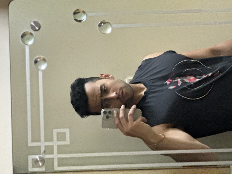 A man takes a selfie in a mirror. He holds his phone with his right hand, showing off a gold bracelet. He wears a black t-shirt with a circular design featuring red and white accents. The mirror has a beige background with decorative glass balls lining the top and left side, connected by linear white designs. The man has dark hair and a serious expression.