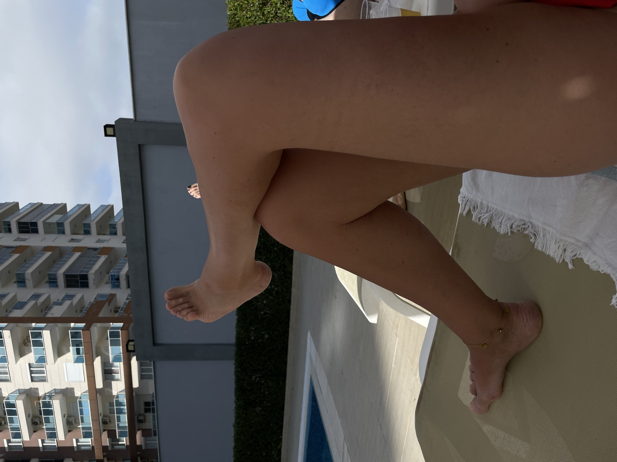 A vertical, full shot depicts a woman's legs crossed on a deck chair, appearing in an outdoor setting next to a pool. The woman is only partially visible, with focus on her bare legs and feet. One foot is adorned with a gold anklet.
The background features a tall building with multiple balconies and windows, indicating an urban setting. A gray wall and a strip of green hedge are also visible. The sky is overcast.
The location appears to be a poolside deck within a city, but the specific city is not apparent from the image.