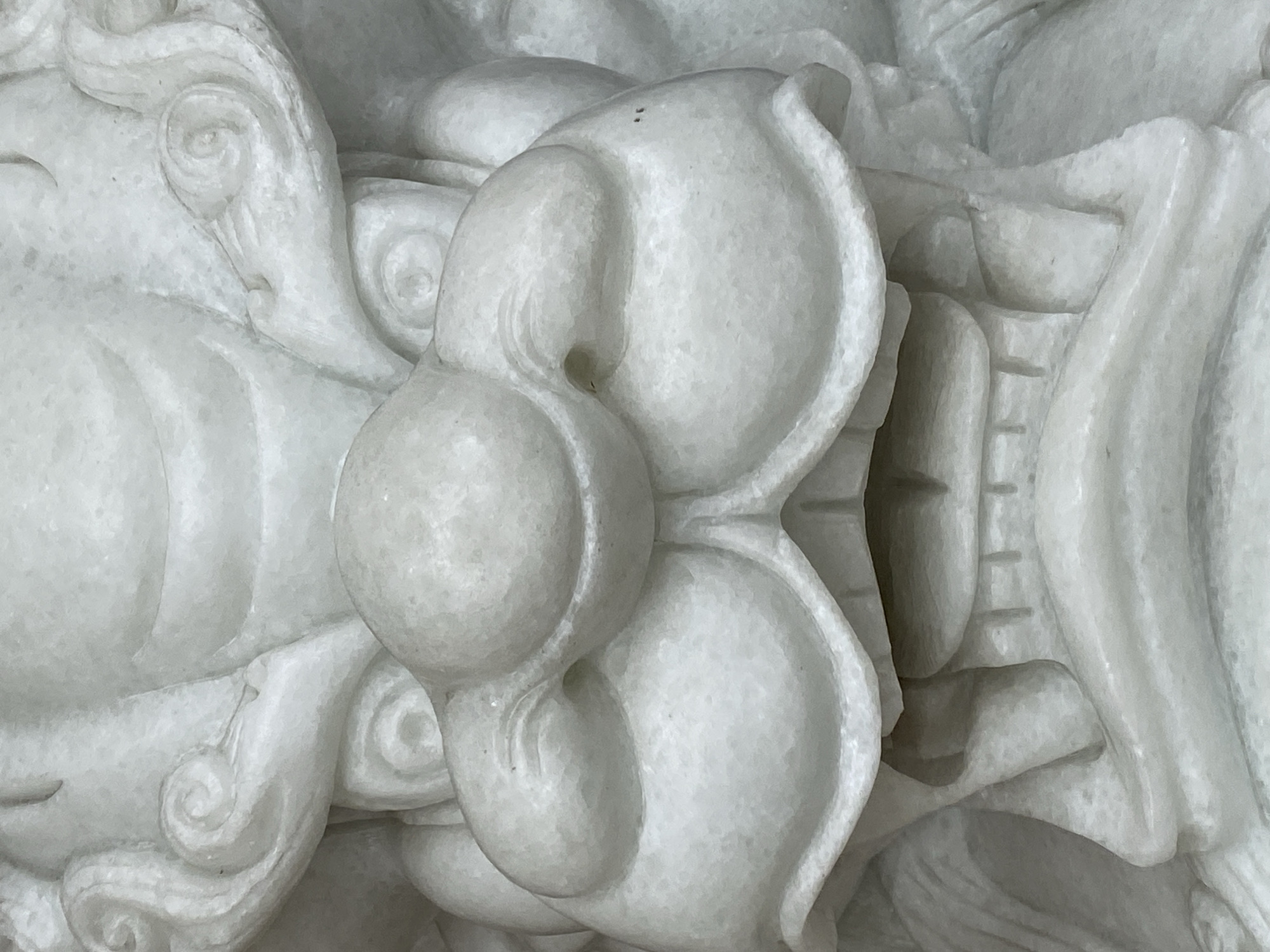 The image shows a close-up, head-on view of a white marble sculpture of a mythical creature, likely a dragon or a lion, with a fierce expression. The sculpture features prominent, bulging eyes, a broad nose with flared nostrils, and an open mouth revealing carved teeth. Swirling patterns adorn the creature's head, possibly representing mane or clouds. The material appears to be marble, with visible veins and a slightly rough texture. The sculpture is likely a decorative element found in a temple, palace, or public space. There is no discernible location in Da Lat, Vietnam depicted in this image.