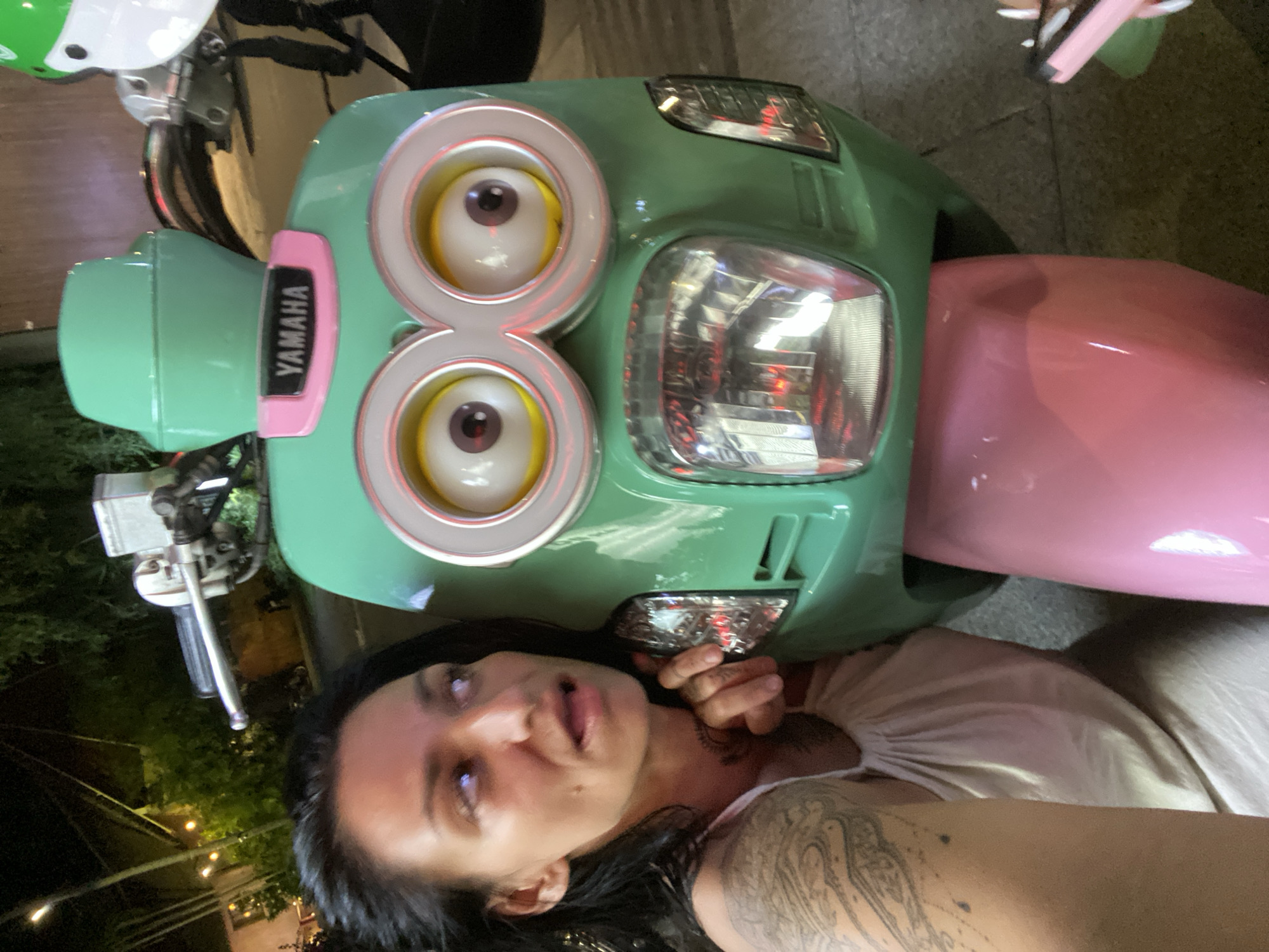 A woman with dark hair and tattoos on her arm poses for a selfie with a green Yamaha scooter modified to resemble a Minion character from the Despicable Me franchise. The scooter features large, cartoonish eyes and a pink front panel. The woman is wearing a white tank top. The background shows a dimly lit urban environment with trees and some lights, suggesting it is nighttime. The scooter's location is Phu Thuy Ward, Vietnam.