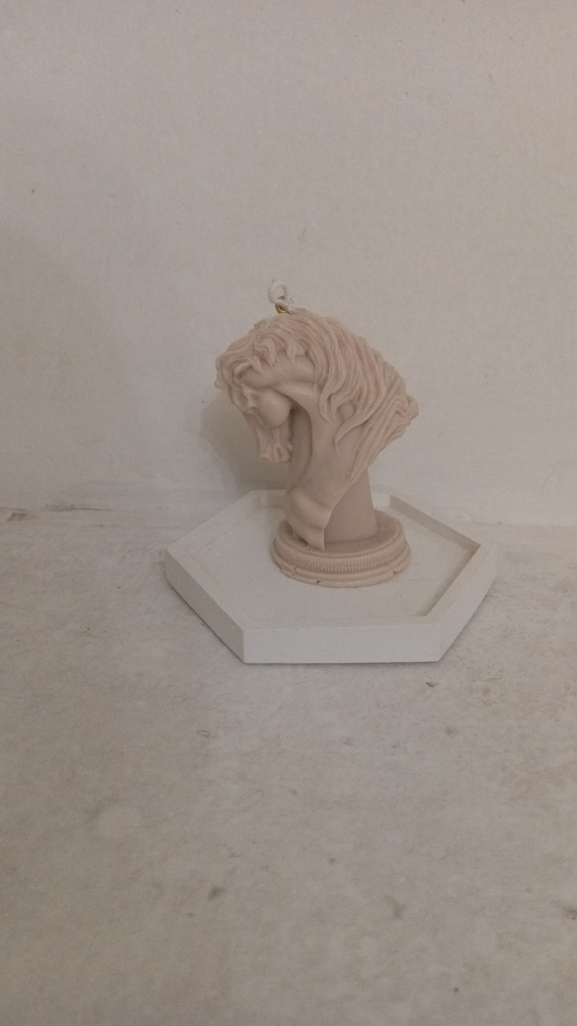 A pale pink, stylized horse head sculpture sits on a white, hexagonal tray. The sculpture is positioned on the right side of the image, facing left and slightly downwards, with its mane flowing. A small loop of string or wire is attached to the top of the sculpture. The tray is placed on a textured, light-colored surface. The background is a plain, off-white wall. The image does not contain any information about the city of Ashtarak, Armenia.
