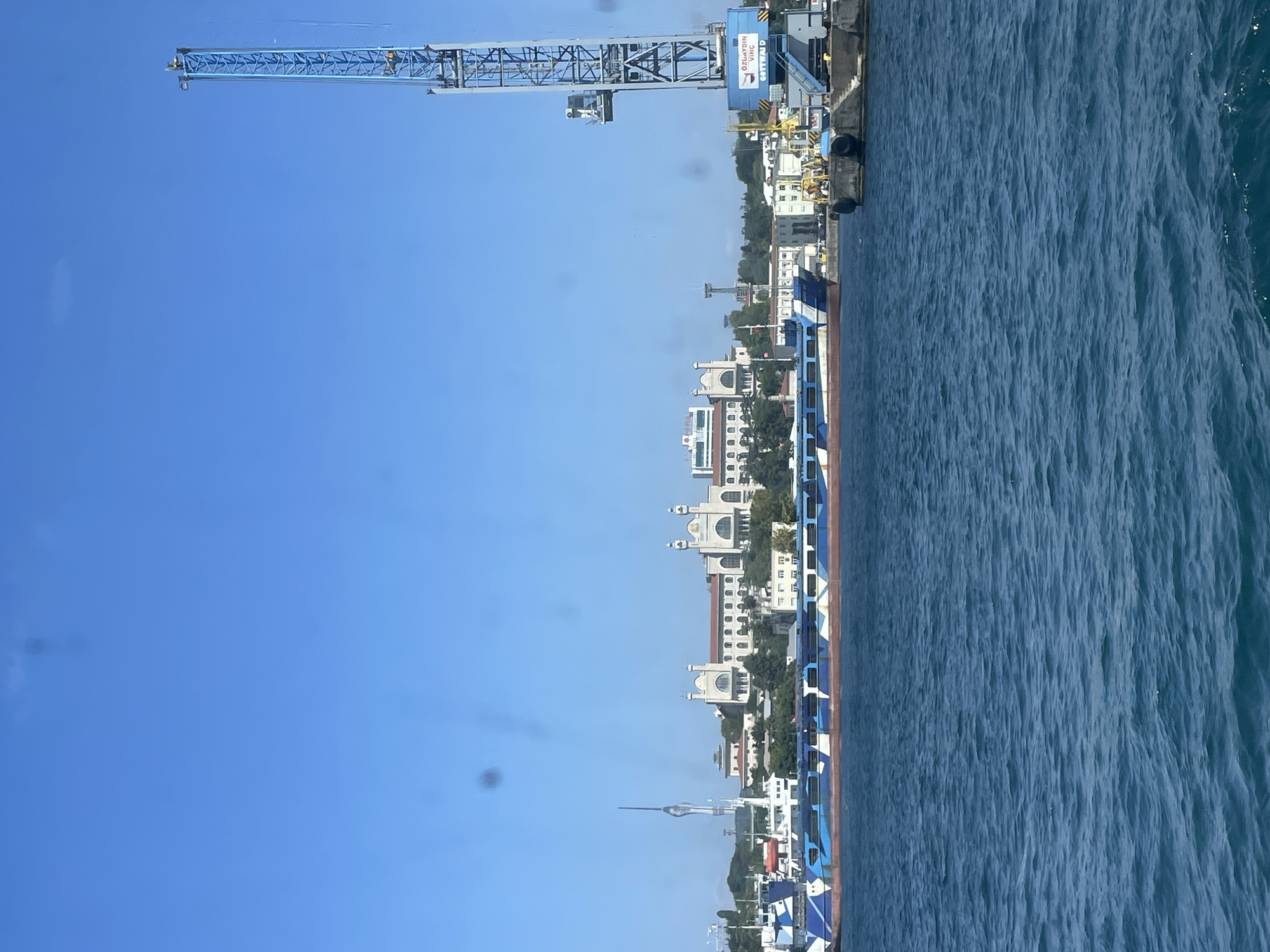 A wide-angle, eye-level, exterior shot captures a bright, sunny day over the waters of the Bosphorus Strait in Istanbul, Turkey. In the foreground, the deep blue water is rippled by a gentle breeze. Dominating the mid-ground is a long, barge-like vessel with a striking blue and white camouflage pattern covering its side. Behind the barge, the Istanbul skyline emerges. Prominent structures include a grand, white building with arched windows and a red-tiled roof, likely a historical institution, with two domes visible. To the left of this building, a slender, modern tower with a distinctive, spire-like top stands tall, contrasting with the older architecture. Further in the background, a variety of other buildings and trees are visible, nestled on the hills overlooking the strait. On the right side of the frame, a large, blue harbor crane with the name "ÖZUAYDIN VINC" is clearly visible, along with associated docking structures and equipment. The sky is a clear, vibrant blue with a few faint smudges or lens flares. There are no discernible people in the image.