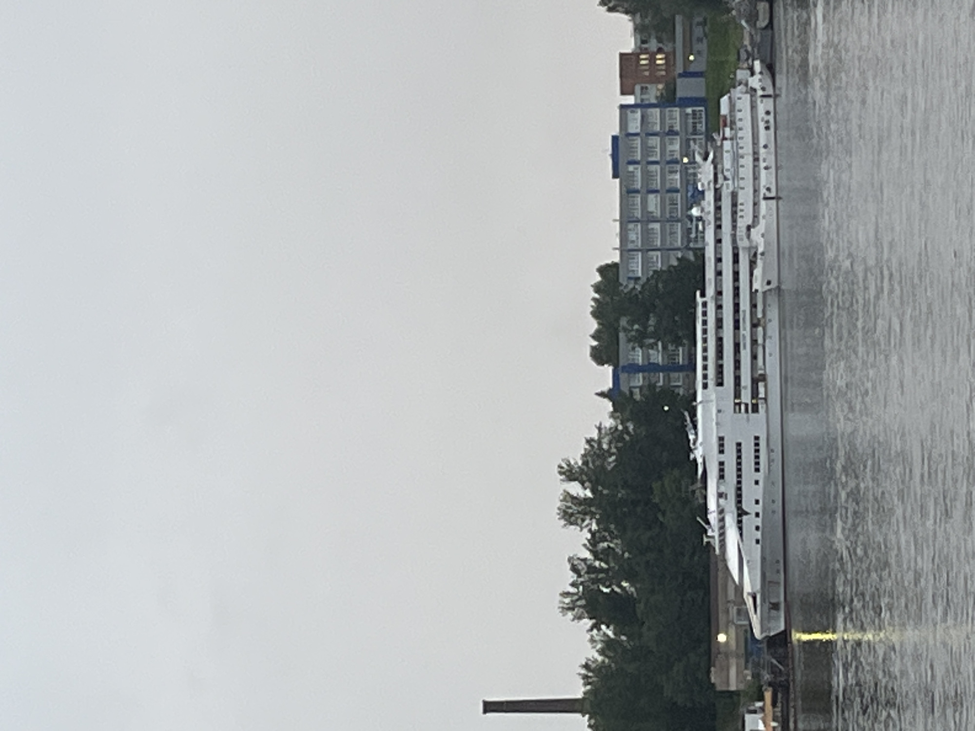 The image shows a large, white cruise ship docked in a body of water. The ship has multiple decks with numerous windows and lifeboats visible. Behind the ship, there is a line of lush green trees. Further in the background, a multi-story building complex with blue trim and rows of windows is visible. To the left of the trees, a tall, dark brick chimney stands against a cloudy, overcast sky. The water in the foreground reflects the overcast sky and the ship. The scene depicts a quiet, possibly industrial, waterfront area in the city of Imeni Sverdlova, Russia.