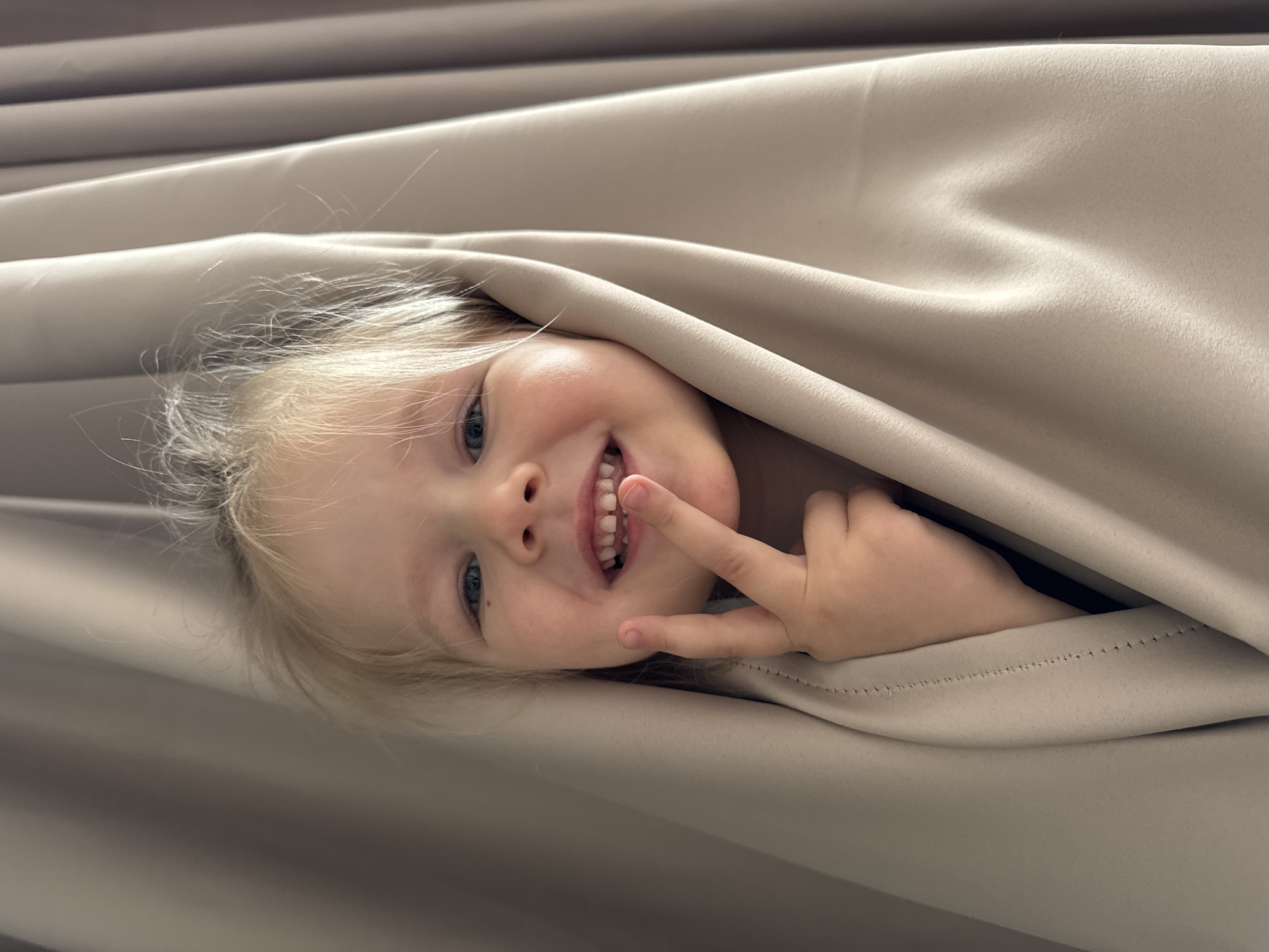 A child with blonde hair is peering out from under a light beige curtain. The child is smiling and has one finger placed near their mouth. The background consists primarily of the draped curtain. It is impossible to identify the location in the city of Gornyi (Dvurechenskoe S/P), Russia from this image.