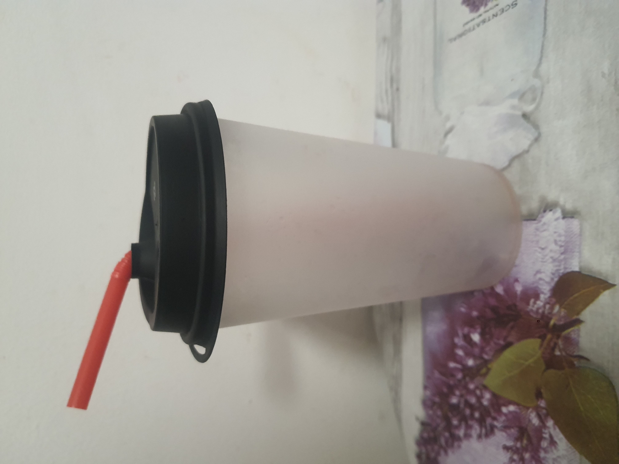 A plastic cup with a black lid and a red straw is shown on a surface with a floral pattern. The cup is translucent and appears to contain a pink beverage. The background is a plain, lightly textured wall. The lighting is diffuse, casting soft shadows. There are no people or animals visible in the image. The overall setting is indoors, likely on a table or counter. The image is taken from a slightly elevated angle, looking down at the cup.