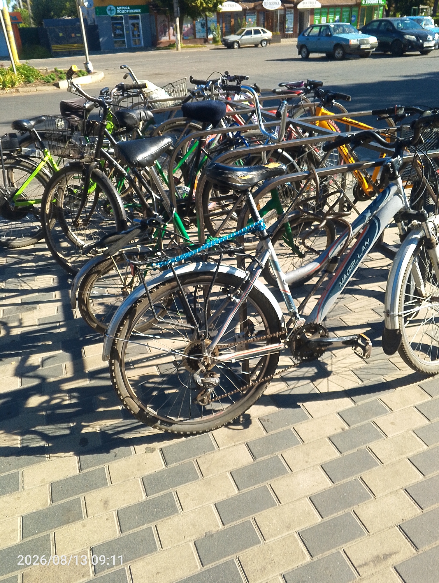 The image shows a row of parked bicycles in front of a store on August 13, 2026, at 09:11. The bicycles are lined up along a sidewalk made of interlocking bricks. The scene appears to be in the city of Bahacheve, Ukraine, as indicated by the text "2026/08/13 09:11" which is likely a date and time stamp, and the presence of Cyrillic text visible on a building in the background which appears to be a pharmacy ("АПТЕКА ПОДОРОЖНИК"). Several cars are visible on the street behind the bicycles. The lighting suggests it is a sunny day.